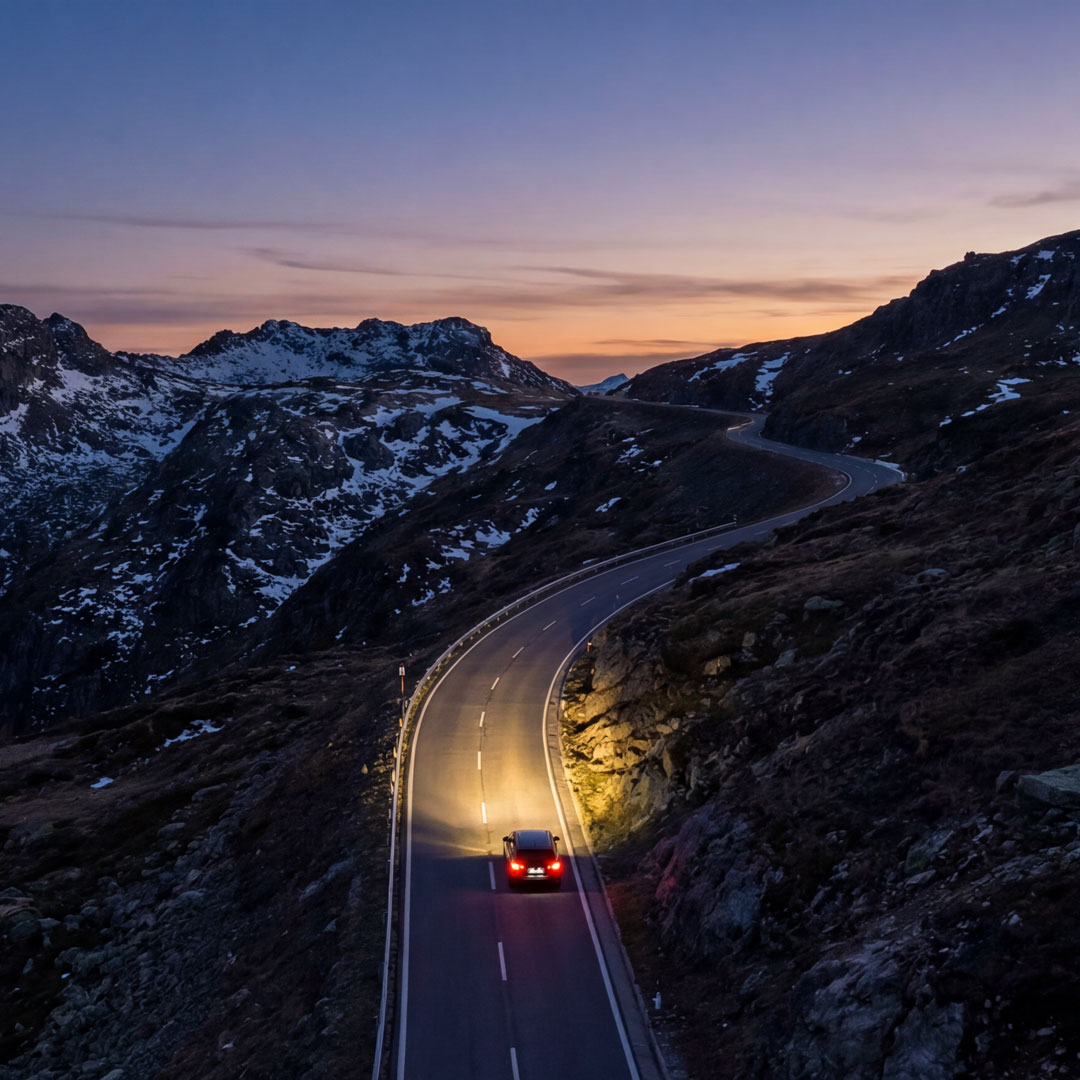Ein Auto fährt bei Dämmerung mit eingeschalteten Scheinwerfern eine kurvige Passstrasse in einer verschneiten Gebirgslandschaft hinauf. Der Himmel leuchtet in sanften Orange- und Blautönen.