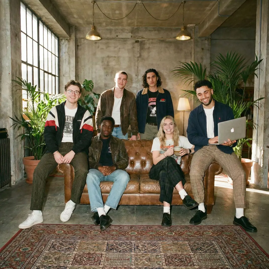 Ein junges, sechsköpfiges Team posiert in einem modern eingerichteten Loft-Studio mit Betonwänden und vielen Pflanzen. Einige sitzen auf einem braunen Ledersofa, während andere dahinter stehen; ein Mann hält einen geöffneten Laptop.