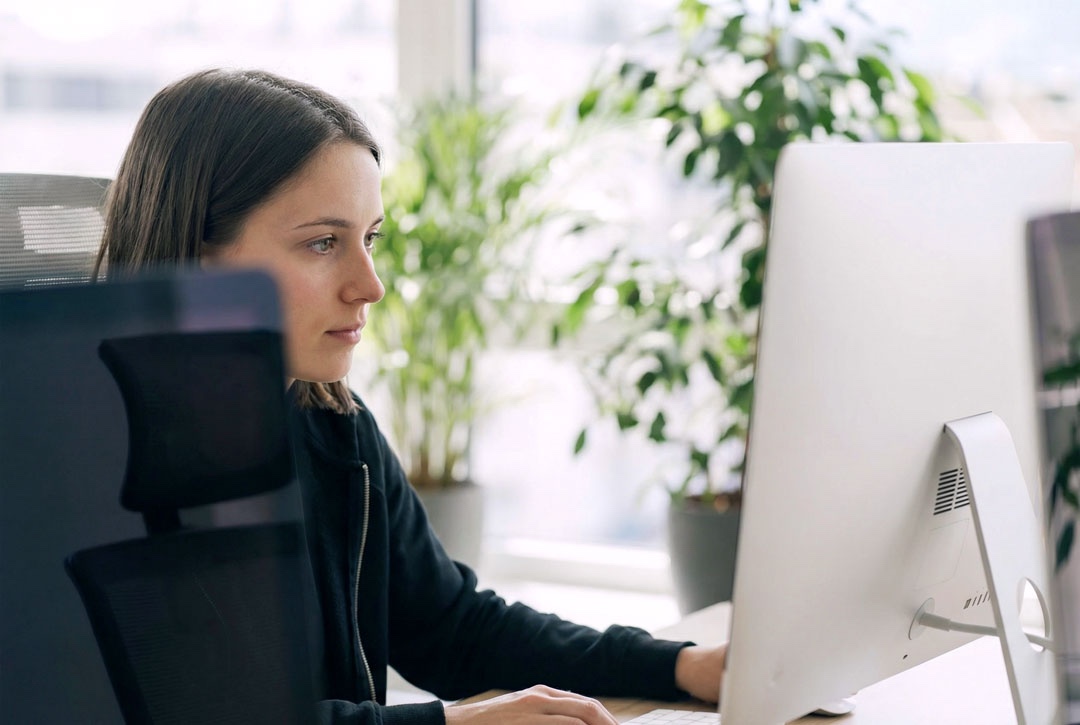 Seitenansicht einer jungen Frau, die aufmerksam auf einen grossen Computerbildschirm blickt. Im hellen Hintergrund befinden sich Zimmerpflanzen und ein Fenster.