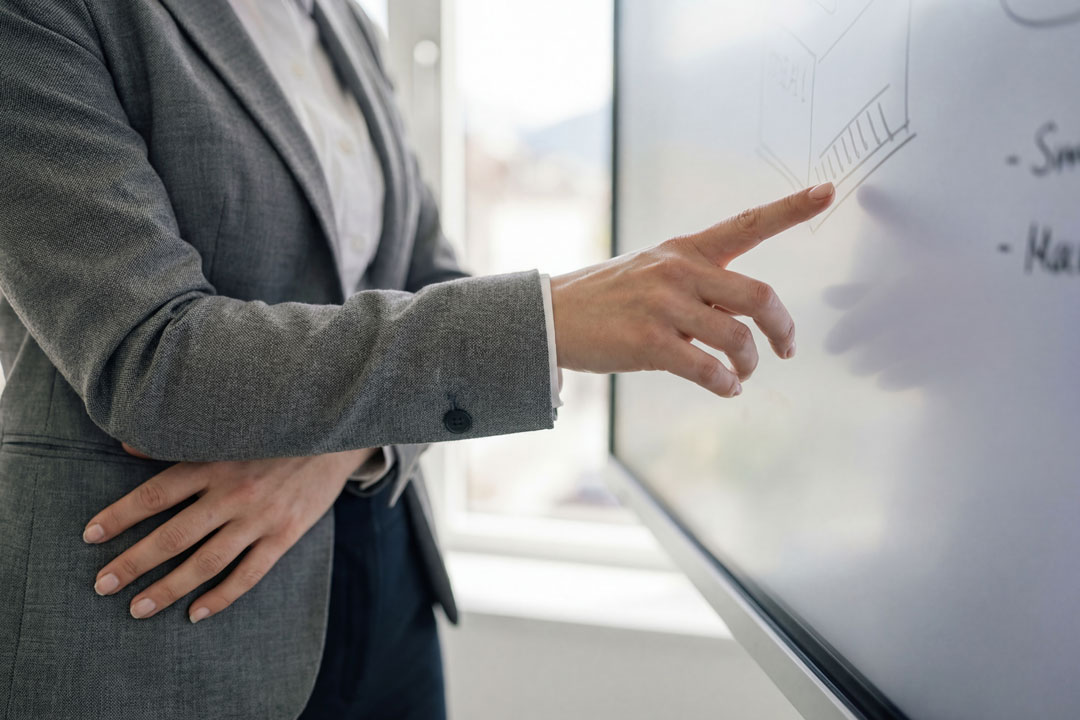 Detailaufnahme einer Person im grauen Blazer, die mit dem Zeigefinger auf eine Skizze auf einem digitalen Whiteboard deutet.