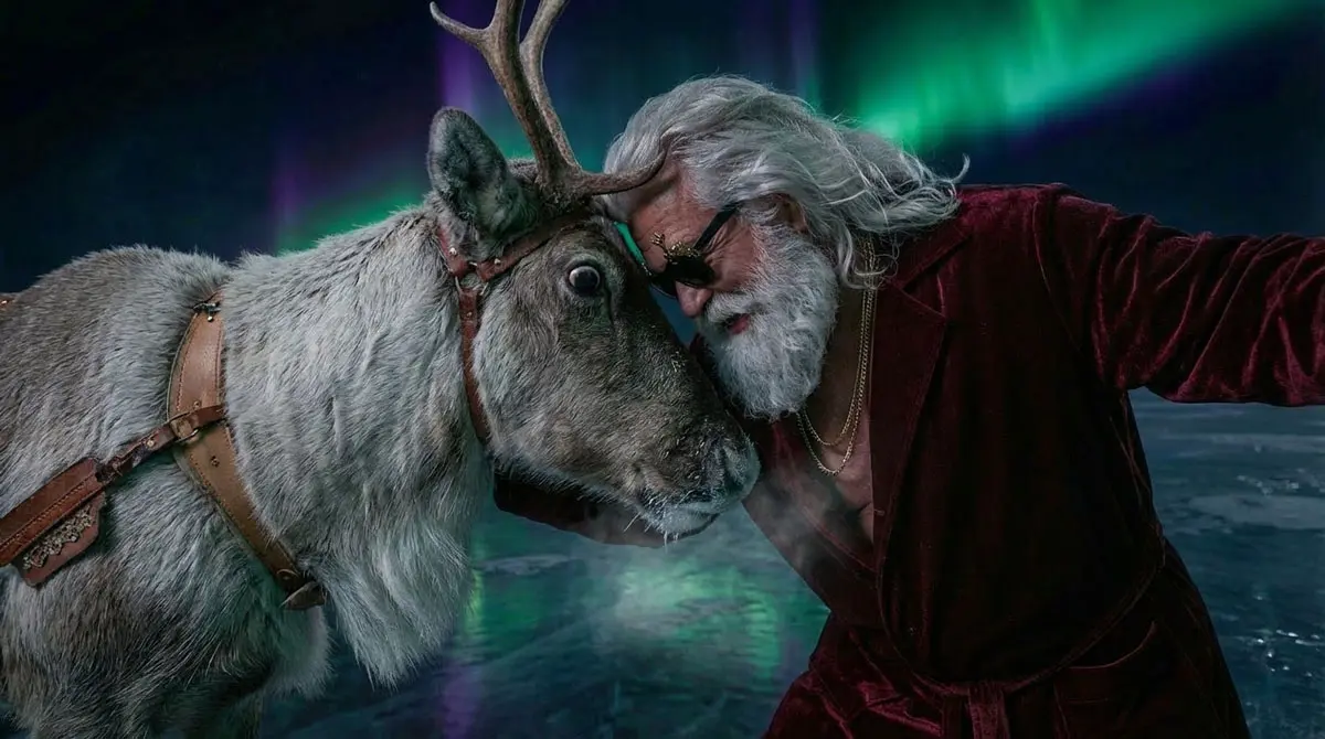 Santa Claus in einem dunkelroten Samtmantel lehnt seine Stirn sanft gegen den Kopf eines Rentiers. Im Hintergrund leuchten grüne und violette Nordlichter über einer vereisten Fläche.