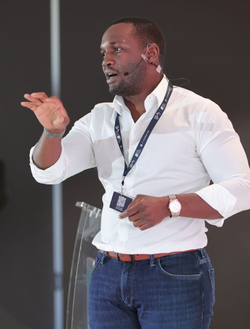 Man in white shirt and jeans speaking with a headset microphone and gesturing with one hand.