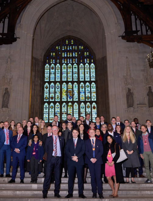 Group of professionally dressed men and women posing on stairs inside a historic building with a large stained glass window in the background.