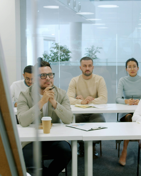 Four adults attentively sitting in a modern conference room with glass walls, listening during a meeting or presentation.