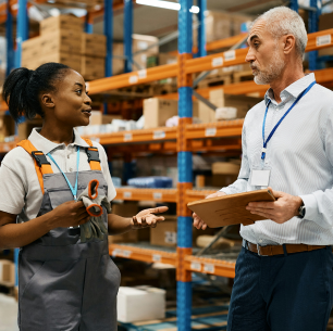 Warehouse worker talking to a supervisor holding a clipboard in an industrial storage area.