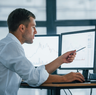 Man in white shirt pointing at financial data on two computer monitors in an office.