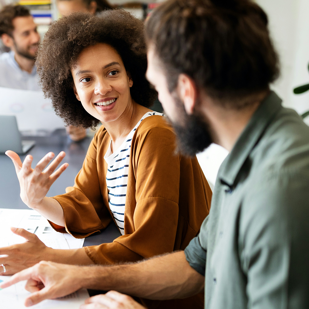 Two colleagues engaged in a discussion over documents in an office setting, with a woman smiling and gesturing.