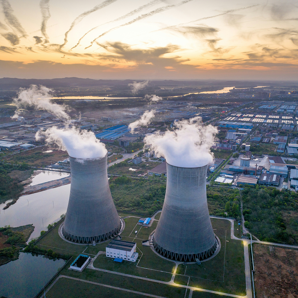 Aerial view of two large cooling towers emitting steam at a power plant during sunset with surrounding industrial buildings and landscape.