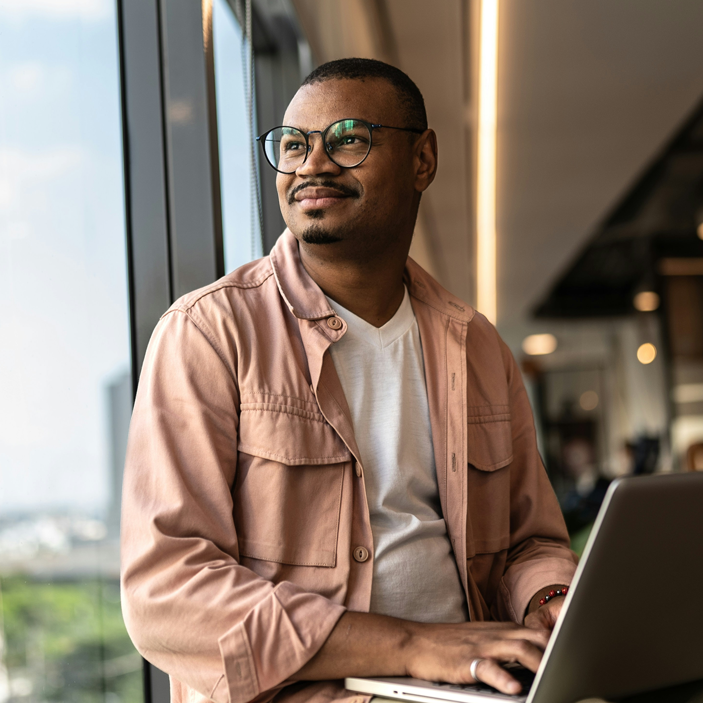 Man wearing glasses and a pink jacket typing on a laptop by a window in an office.