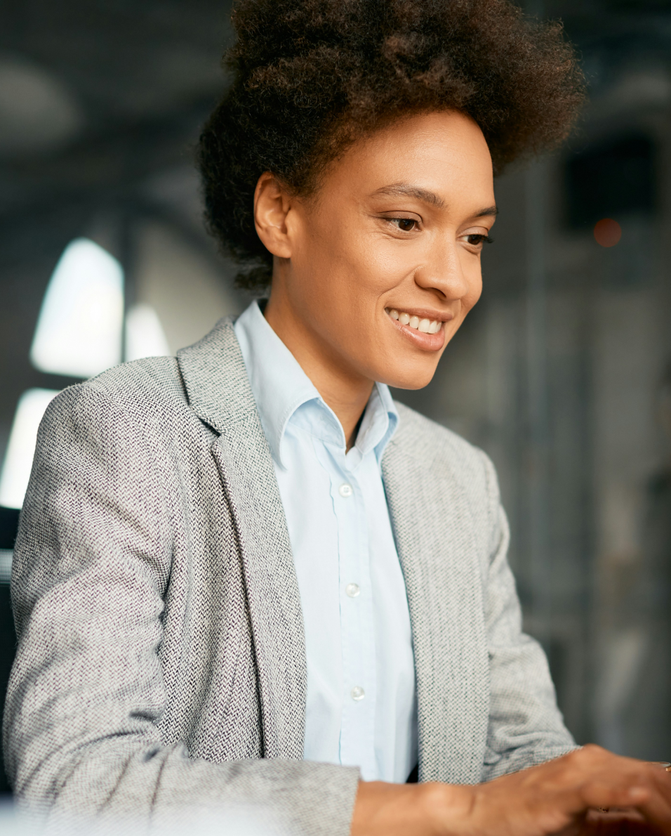Smiling woman with curly hair wearing a gray blazer and blue shirt working at a computer.