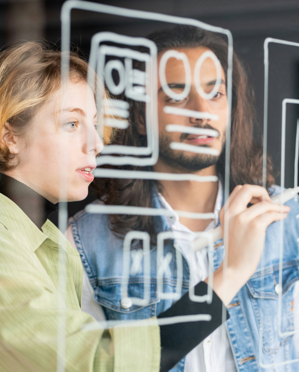 Two people collaboratively drawing a website wireframe on a transparent glass board.