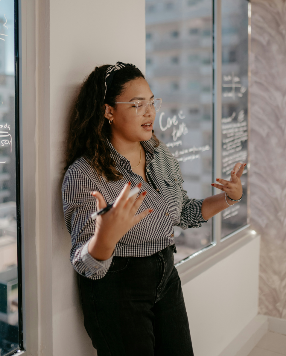 Woman with glasses and curly hair stands by a window covered with writings, gesturing while speaking.