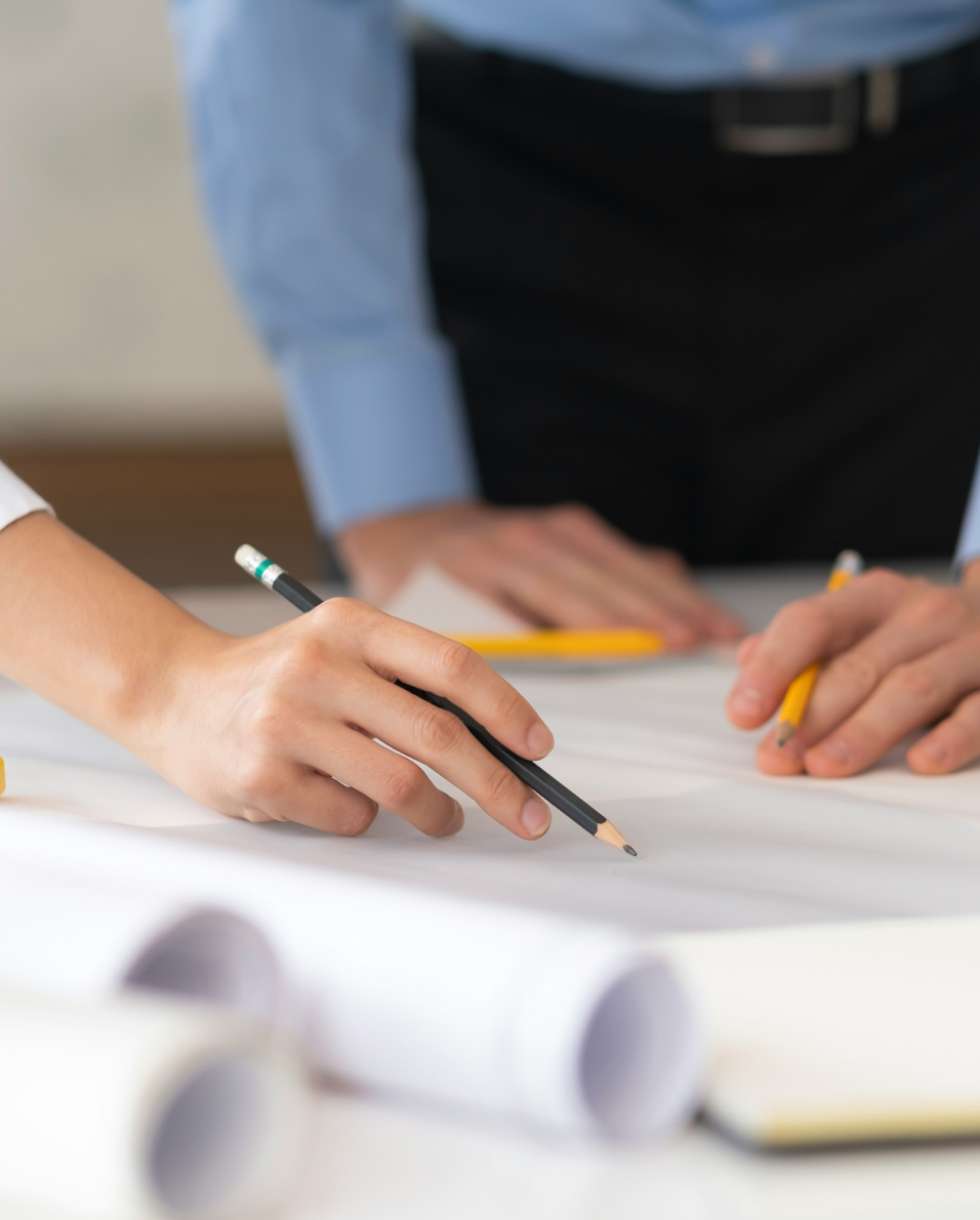 Two people reviewing documents on a table with pencils in hand, with rolled-up papers in the foreground.