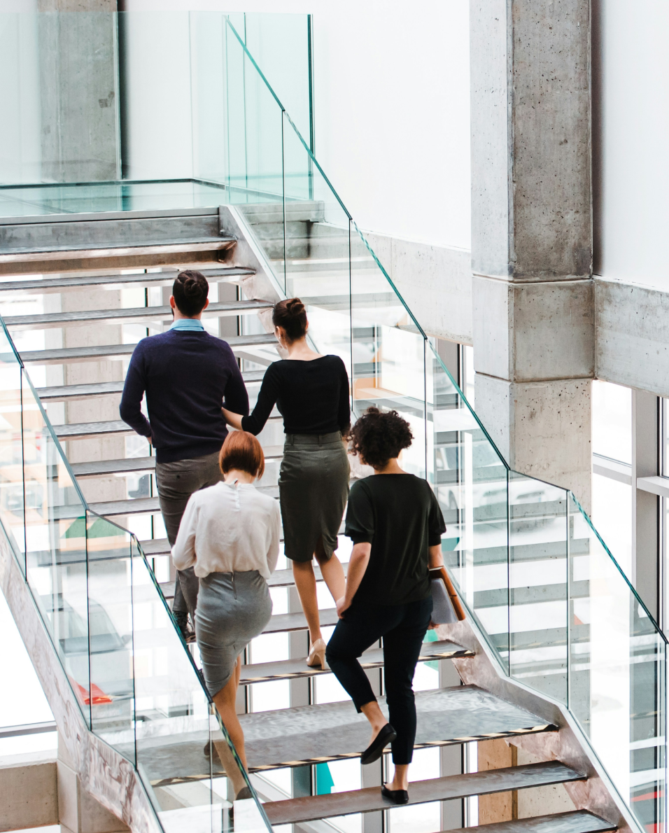 Four business professionals walking up a modern glass and metal staircase inside an office building.