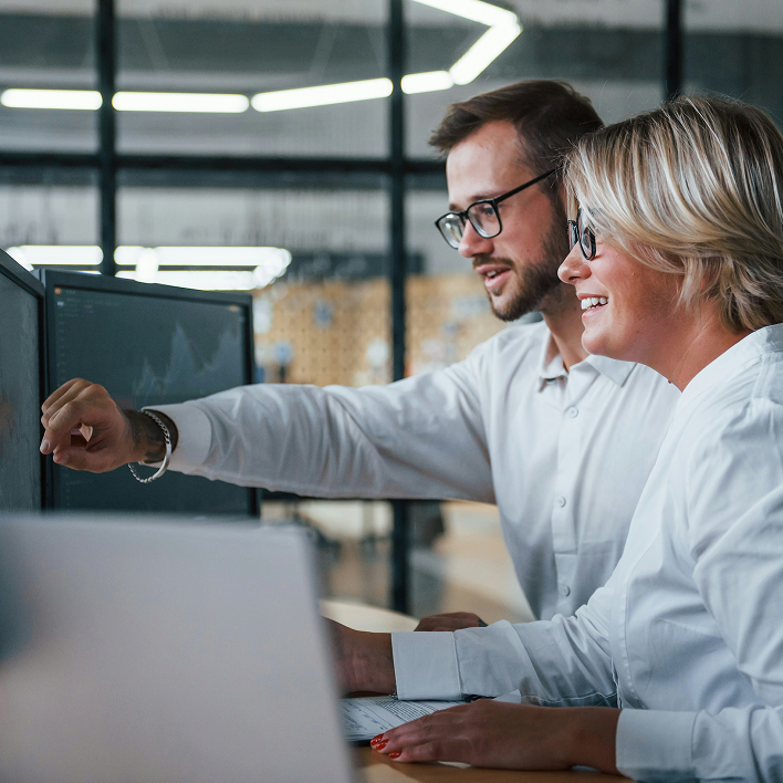 A man and a woman wearing glasses are smiling and looking at a computer screen, with the man pointing at the screen in a modern office setting.