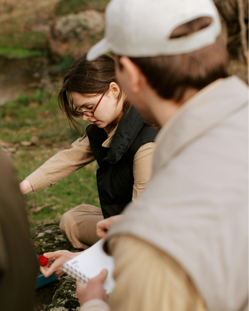 A woman wearing glasses and a black vest kneeling outdoors, while a man in a beige cap and jacket looks on holding a notebook.