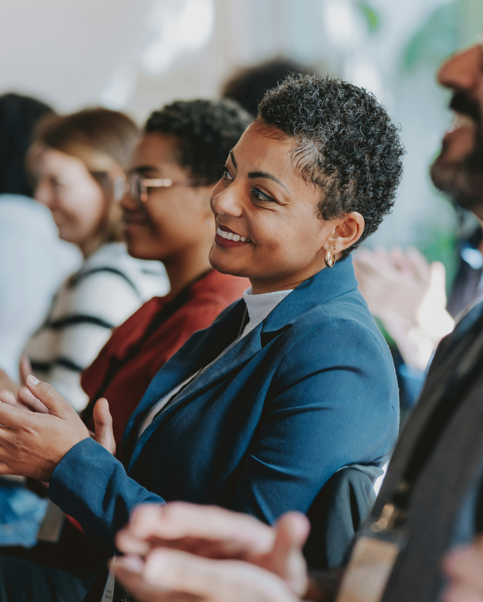 Smiling woman in a blue blazer clapping during a professional event with others in the background.