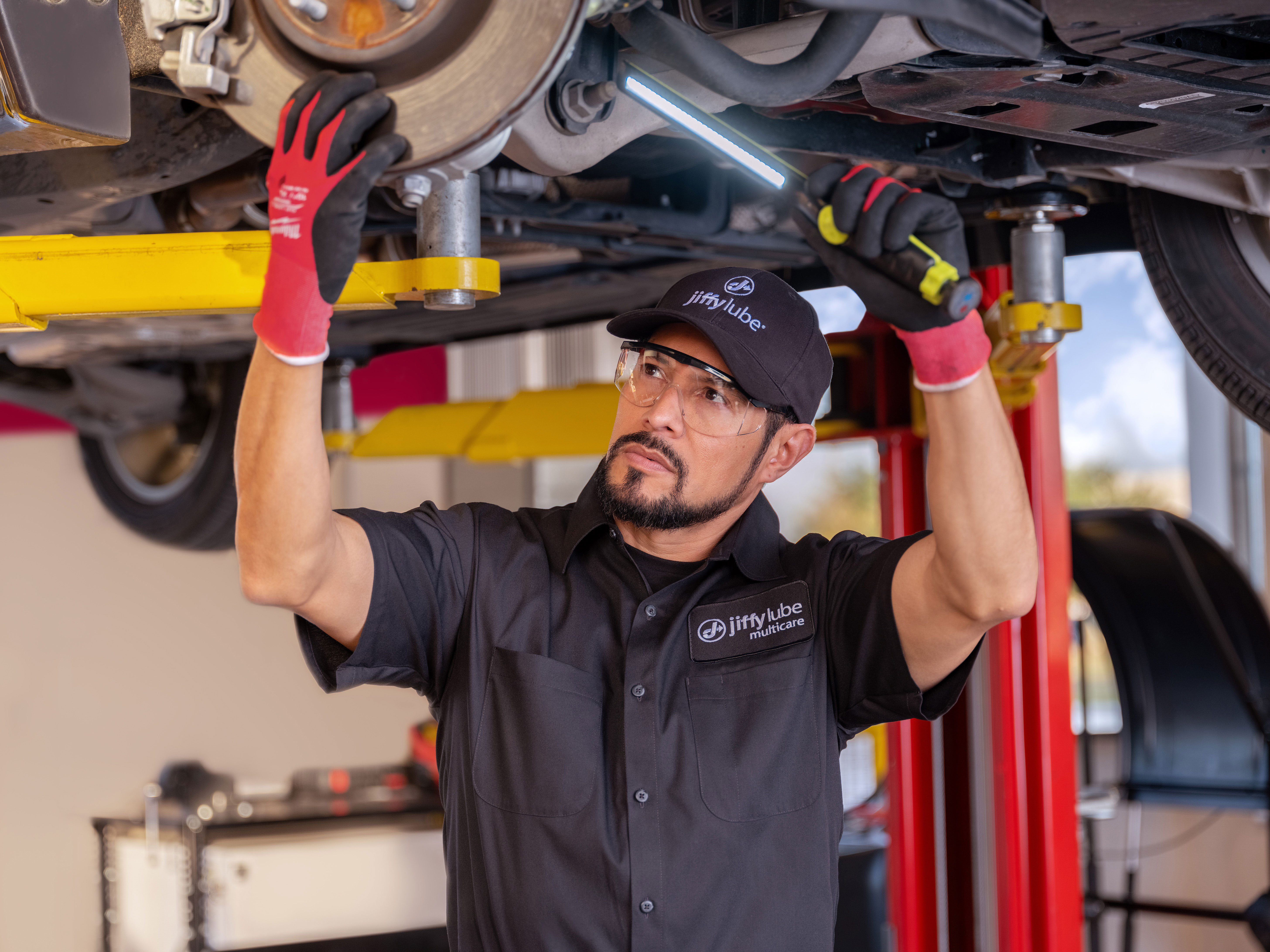 Mechanic in Jiffy Lube uniform inspecting the underside of a vehicle with a flashlight in an auto repair shop.