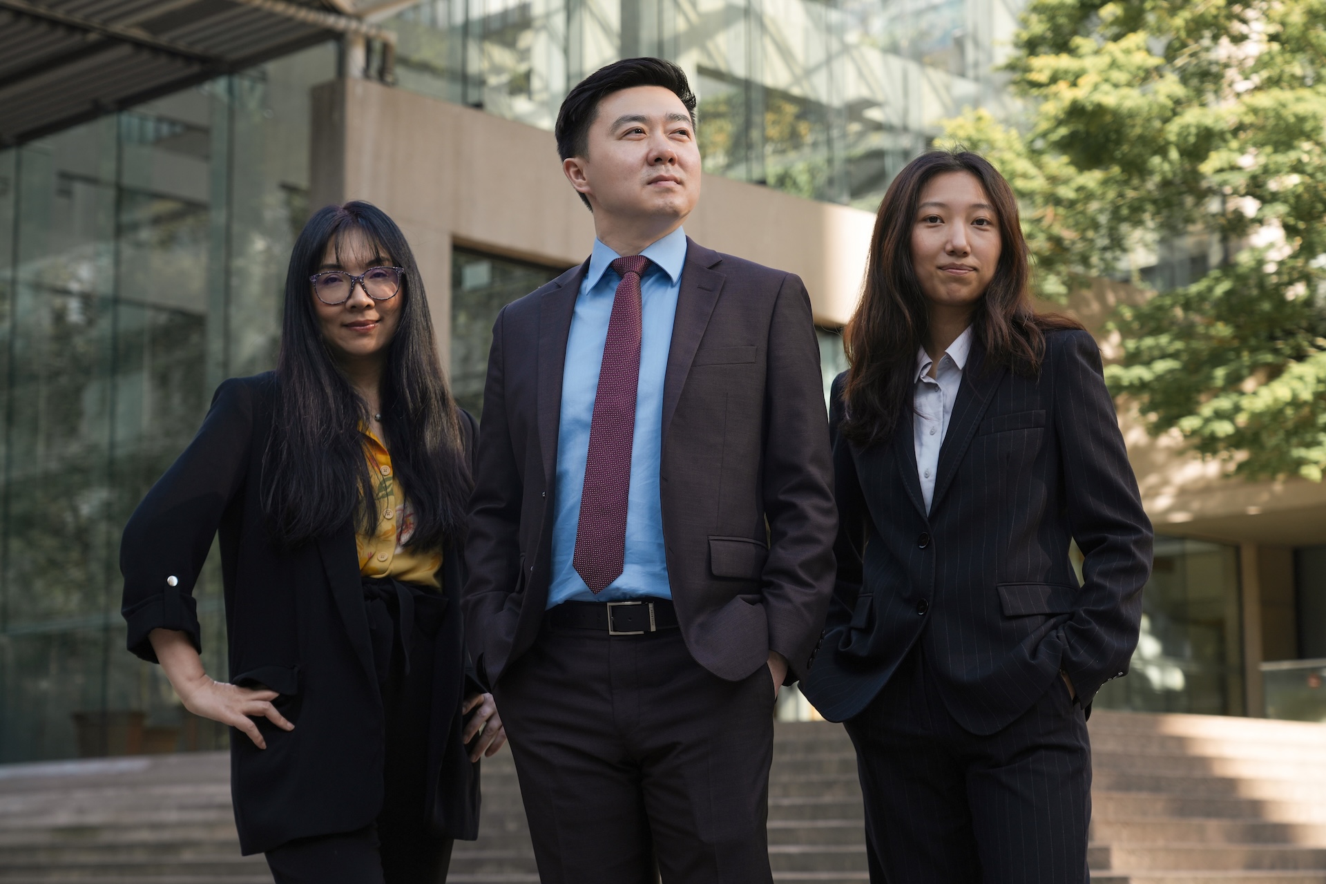 Three confident business professionals standing outside a modern glass building, two women and one man dressed in suits.