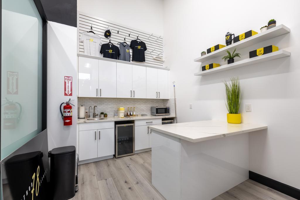 Modern white kitchenette with hanging T-shirts, countertop with sink, small fridge, microwave, and shelves with yellow and black boxes and plants.