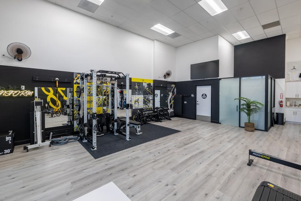 Modern gym with weight rack, dumbbells, resistance machines, and light wood flooring.