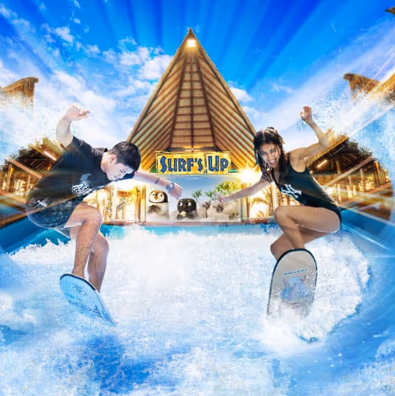 Two surfers performing tricks on waves in front of the Surf’s Up attraction entrance at Columbia Pictures Aquaverse, with a dramatic water splash effect and stylized tropical backdrop.

