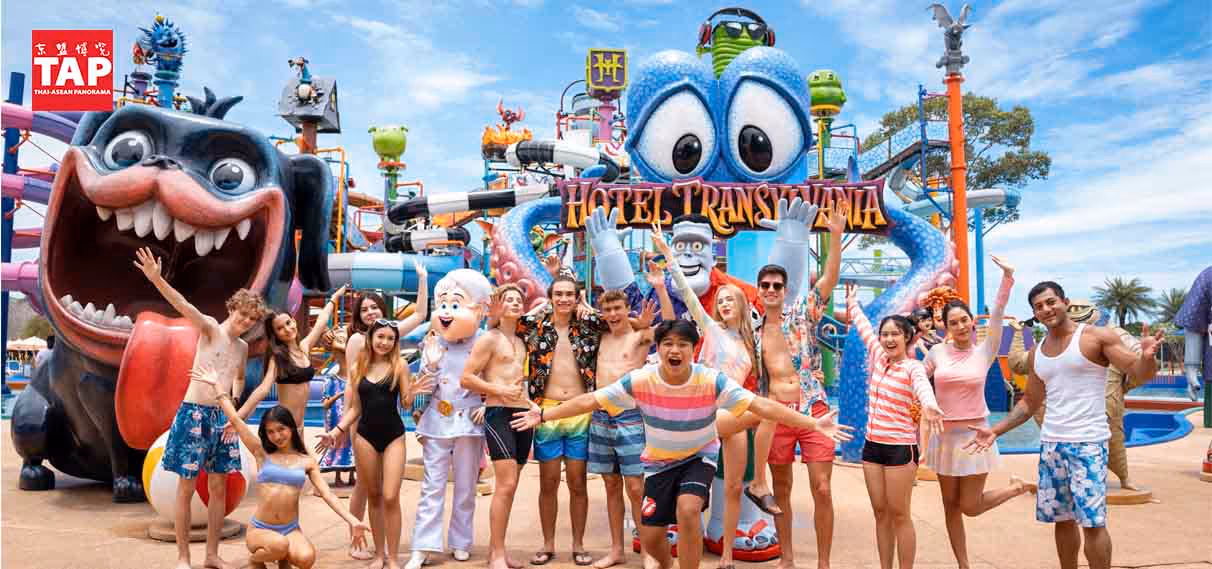 A lively group of young adults in swimwear and summer outfits pose joyfully in front of the Hotel Transylvania themed water play zone at Columbia Pictures Aquaverse. Behind them are colorful water slides, splash features, and large character statues, including a wide-eyed blue octopus and a giant grinning dog. Two mascots from the franchise are also present, adding to the playful atmosphere. The sky is bright blue, and the setting radiates fun, energy, and vacation vibes—ideal for a tropical water park adventure. The TAP (Thai-ASEAN Panorama) logo is visible in the upper left corner.