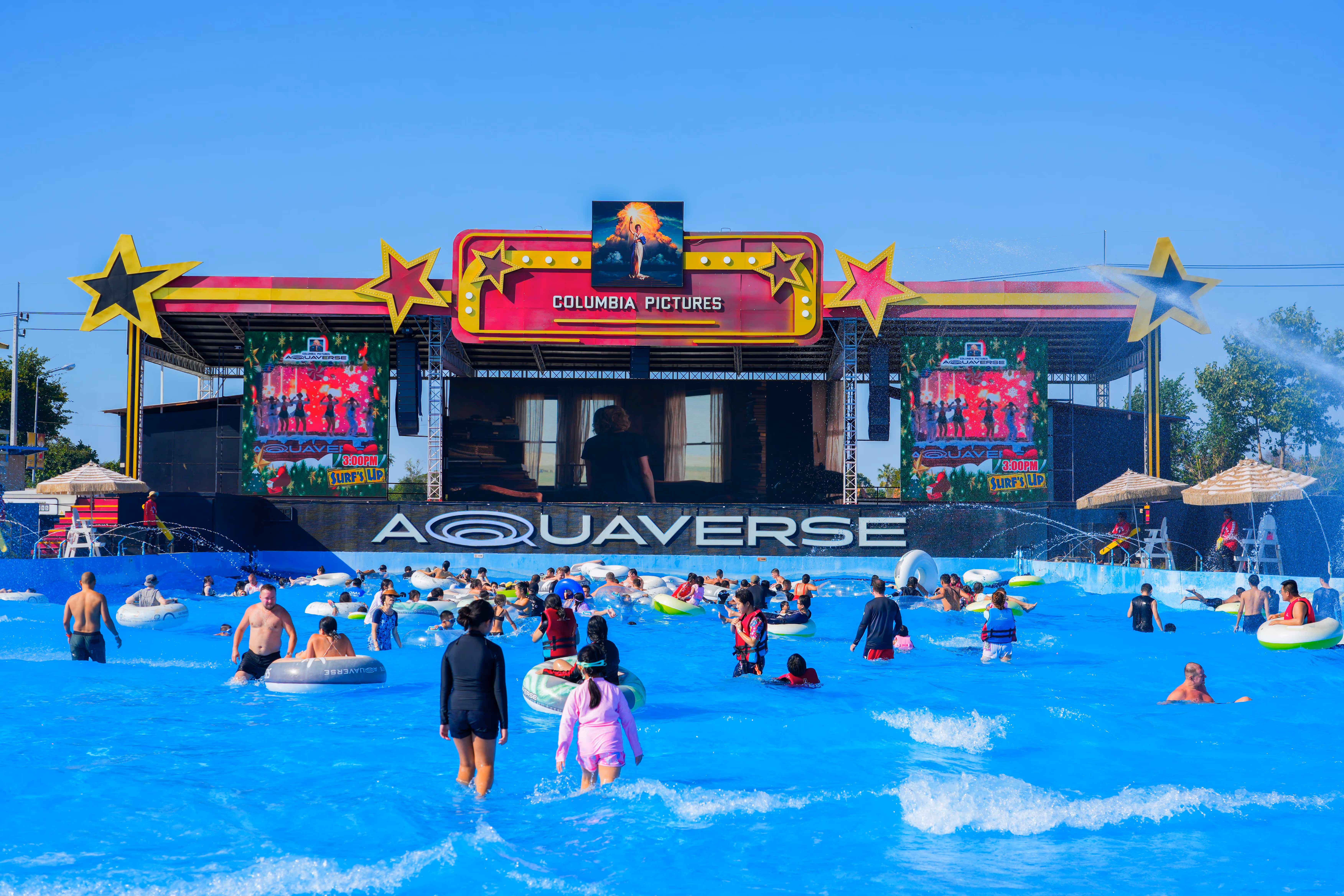 Guests enjoying the vibrant wave pool at Columbia Pictures Aquaverse under a clear blue sky, with a large stage and digital screens playing entertainment content beneath a colorful Columbia Pictures marquee.

