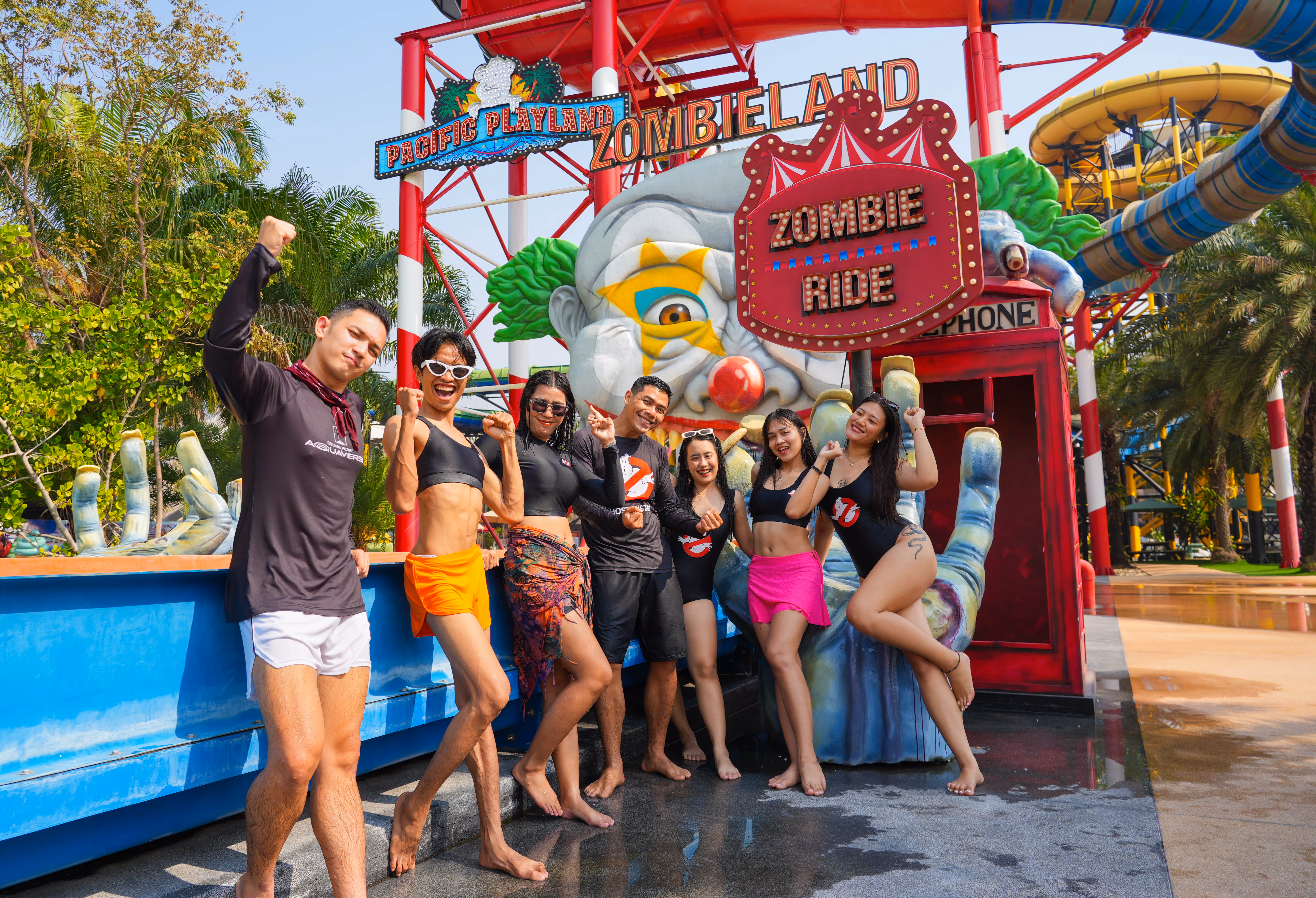 Group of smiling young adults in swimwear posing in front of the colorful 'Zombie Ride' entrance at Columbia Pictures Aquaverse, with a clown face structure and water slides in the background.