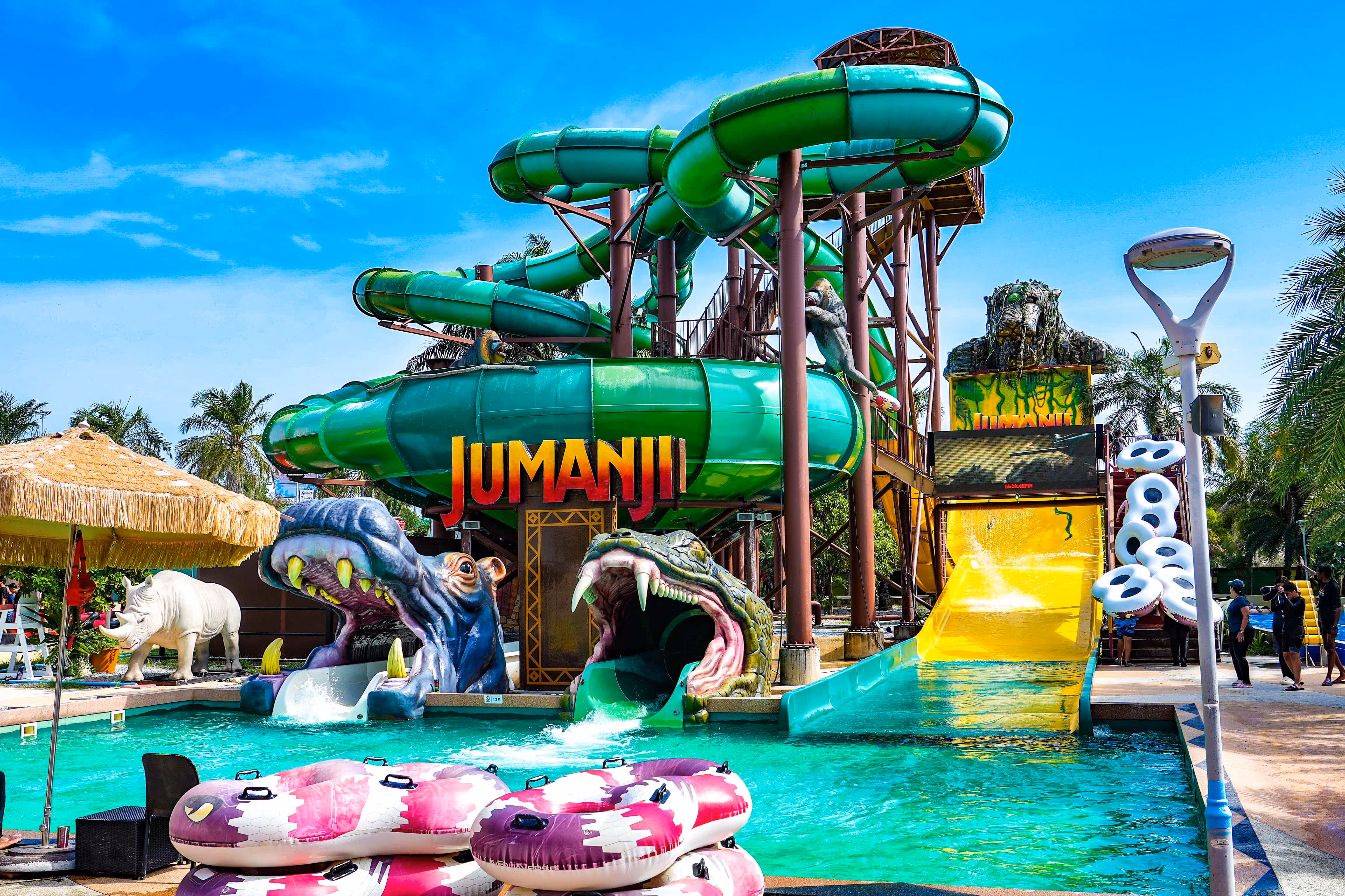 Jumanji-themed water slide attraction at Columbia Pictures Aquaverse, featuring twisting green tube slides, jungle decorations, and a splash pool with pink donut floaties under a sunny blue sky.

