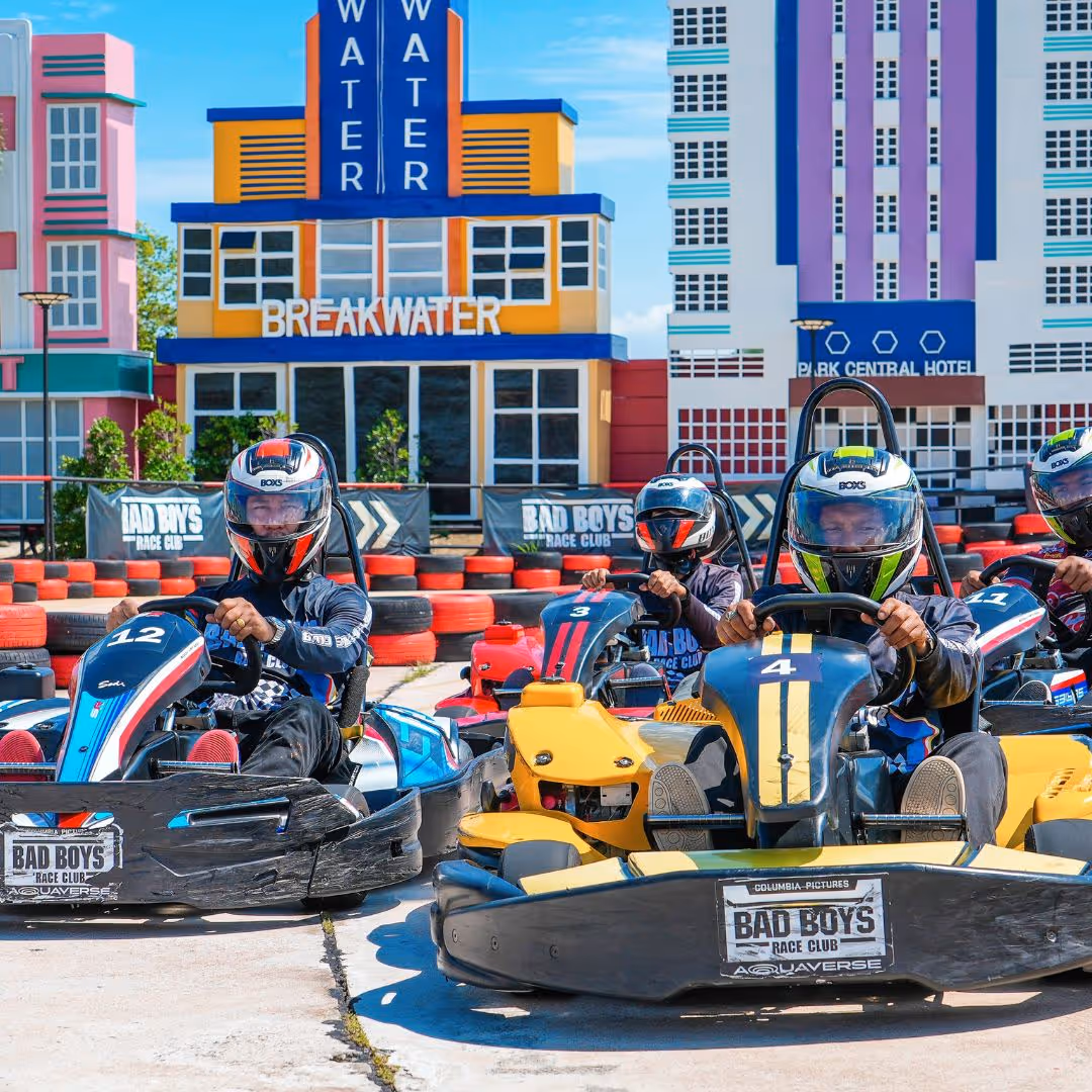 Group of guests in helmets preparing to race go-karts on the Gran Turismo-themed track at Columbia Pictures Aquaverse, with colorful cityscape backdrops and tire barriers surrounding the course.