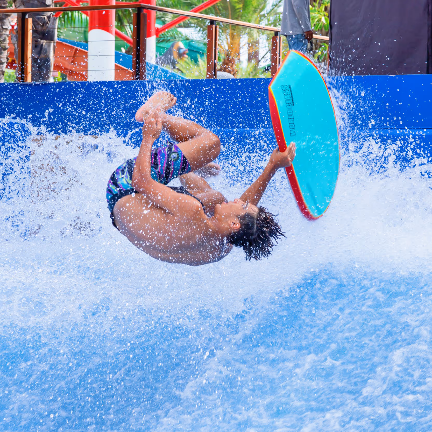 Young man performing a backflip on a bodyboard at the Surf’s Up attraction in Columbia Pictures Aquaverse, surrounded by splashing water and colorful tropical decor in the background.

