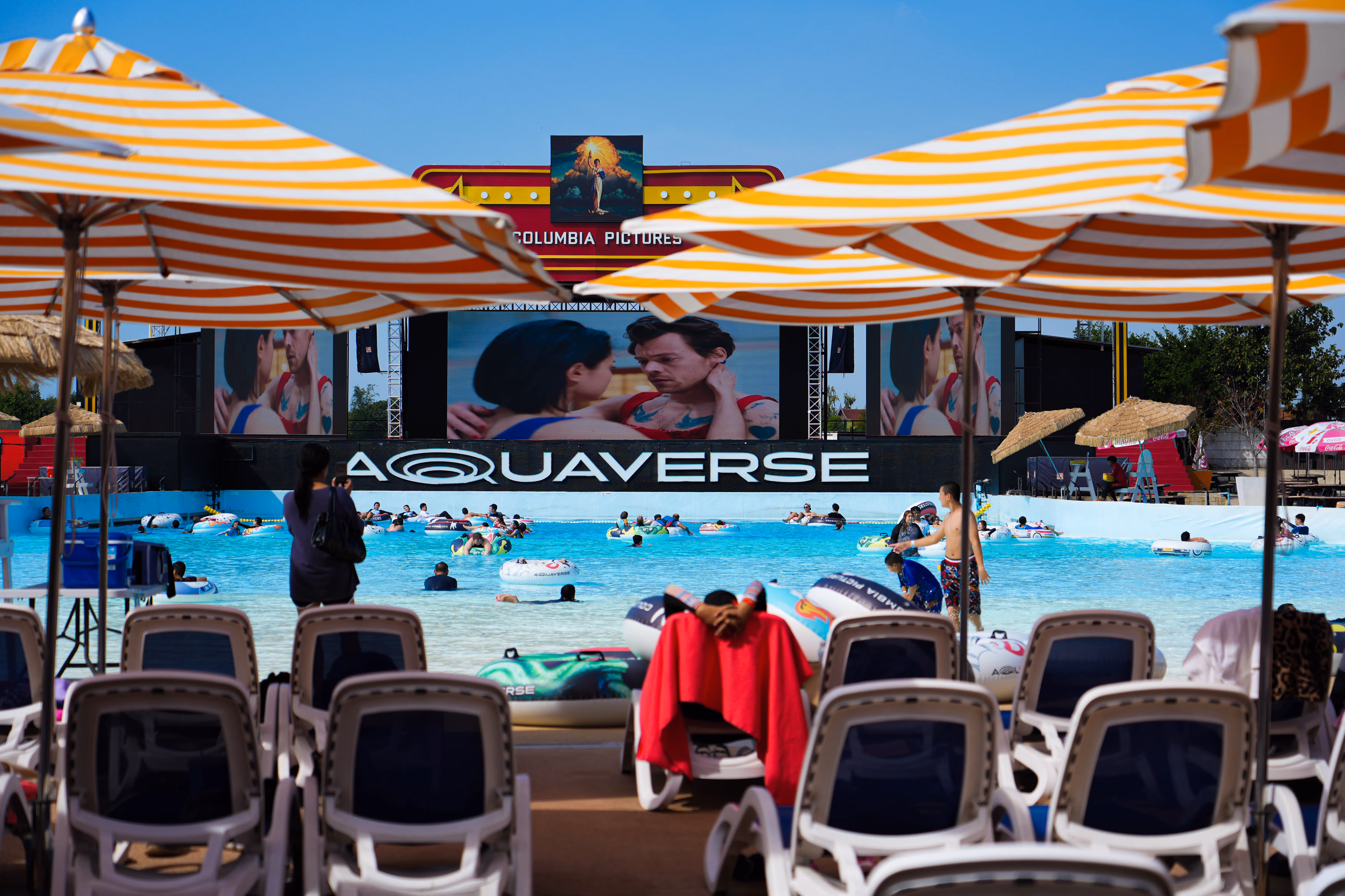 Guests enjoying the vibrant wave pool at Columbia Pictures Aquaverse under a clear blue sky, with a large stage and digital screens playing entertainment content beneath a colorful Columbia Pictures marquee.


