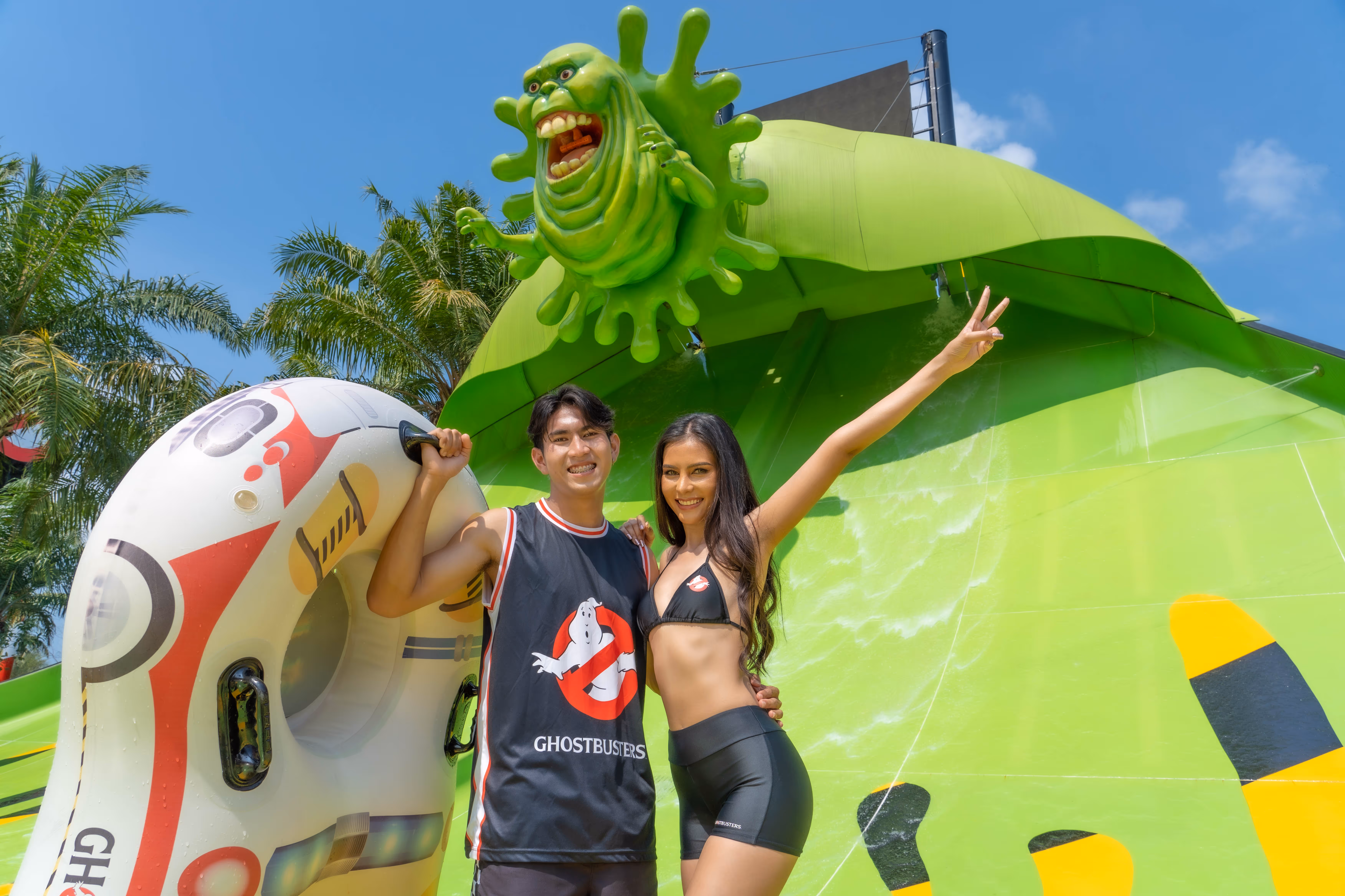 Smiling couple posing in front of a bright green Ghostbusters-themed water slide at Aquaverse, with a large Slimer figure overhead and a themed inner tube in hand.