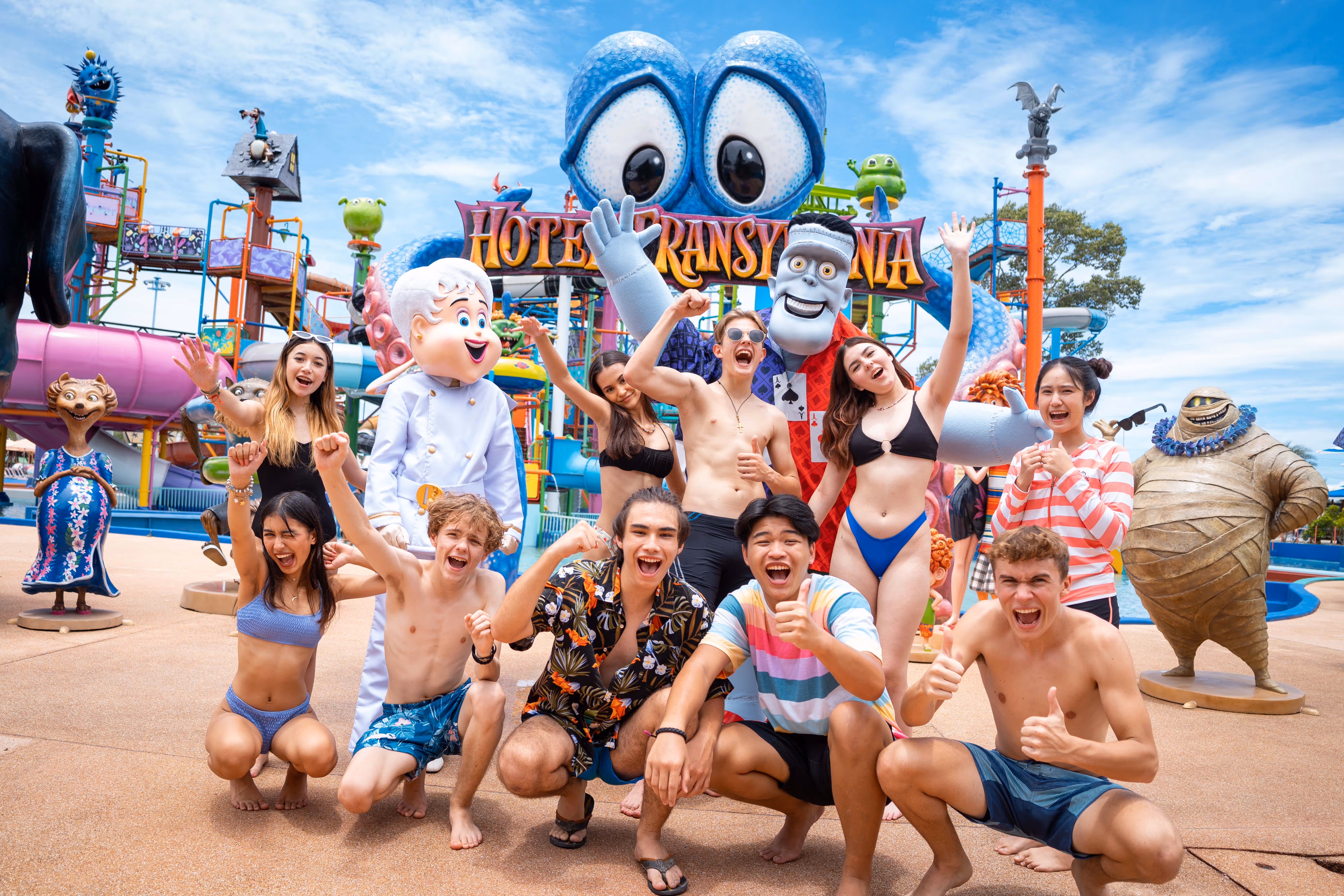 Group of excited guests posing in swimwear at the Hotel Transylvania-themed water play zone in Columbia Pictures Aquaverse, surrounded by life-sized character mascots and colorful splash structures.

