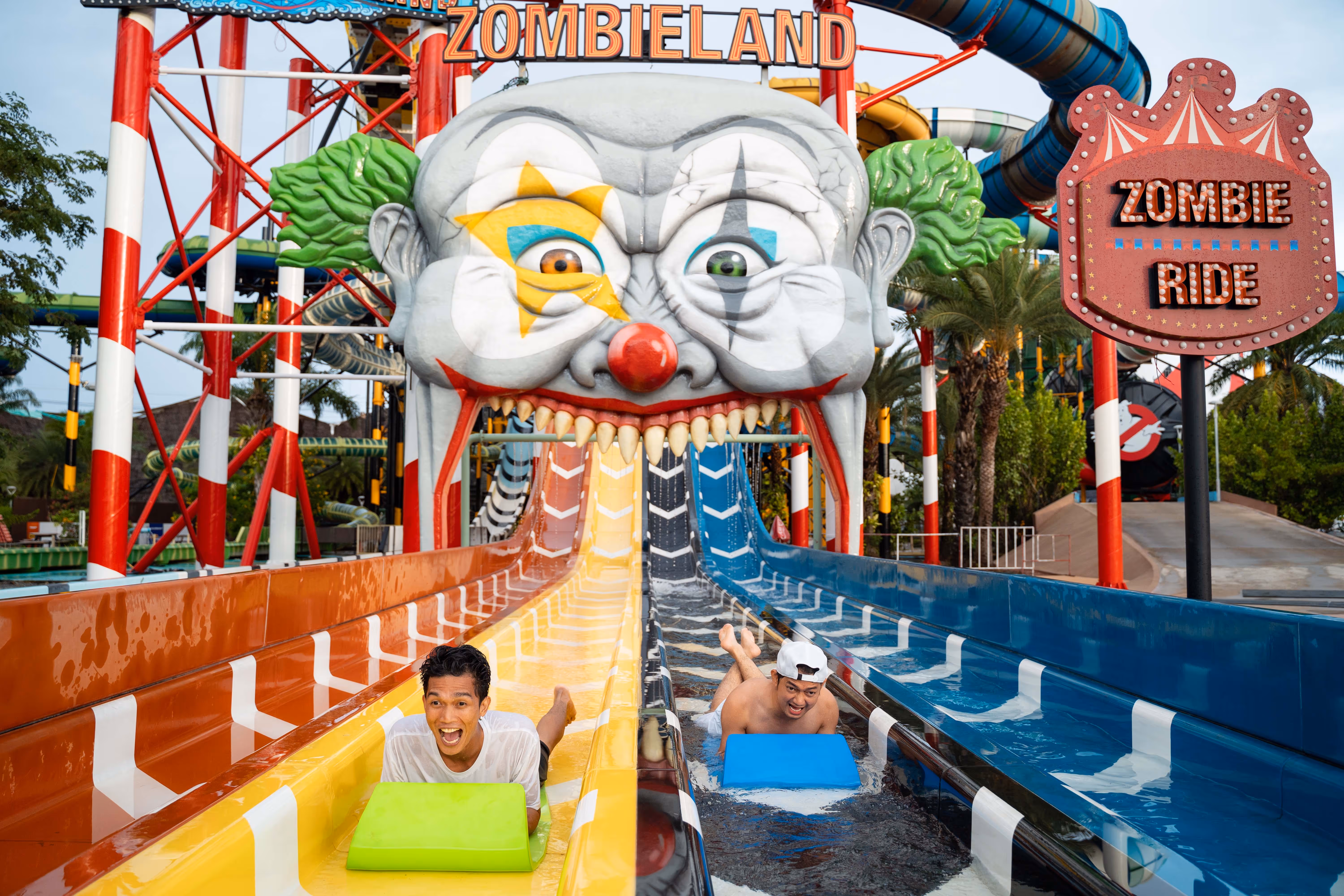 Two guests racing down colorful water slides at the Zombieland-themed attraction in Columbia Pictures Aquaverse, sliding out of a giant creepy clown face with “Zombie Ride” and “Zombieland” signage in the background.