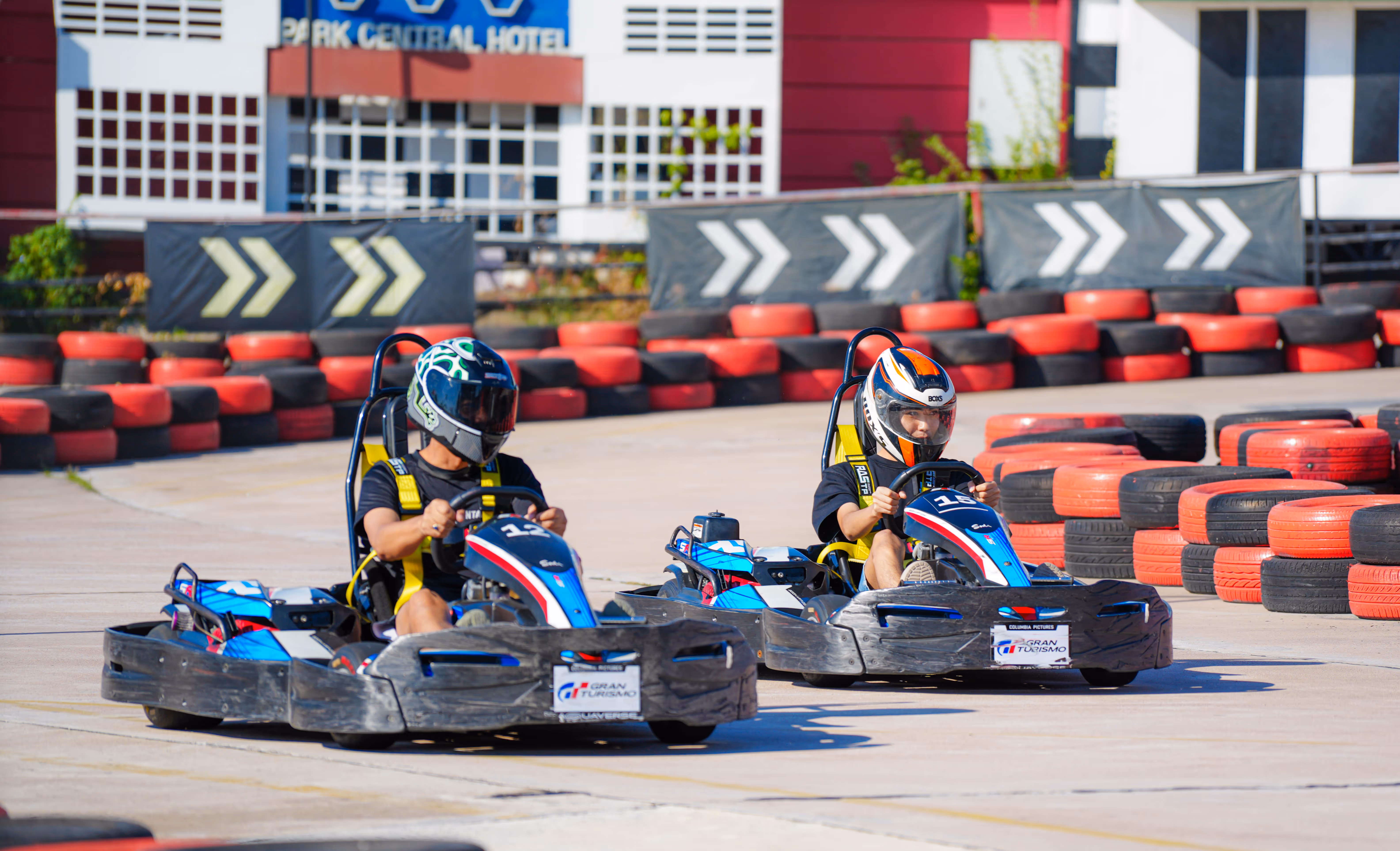 Two guests racing go-karts around a track at Columbia Pictures Aquaverse, wearing helmets and safety gear, with red and black tire barriers and directional signage in the background.

