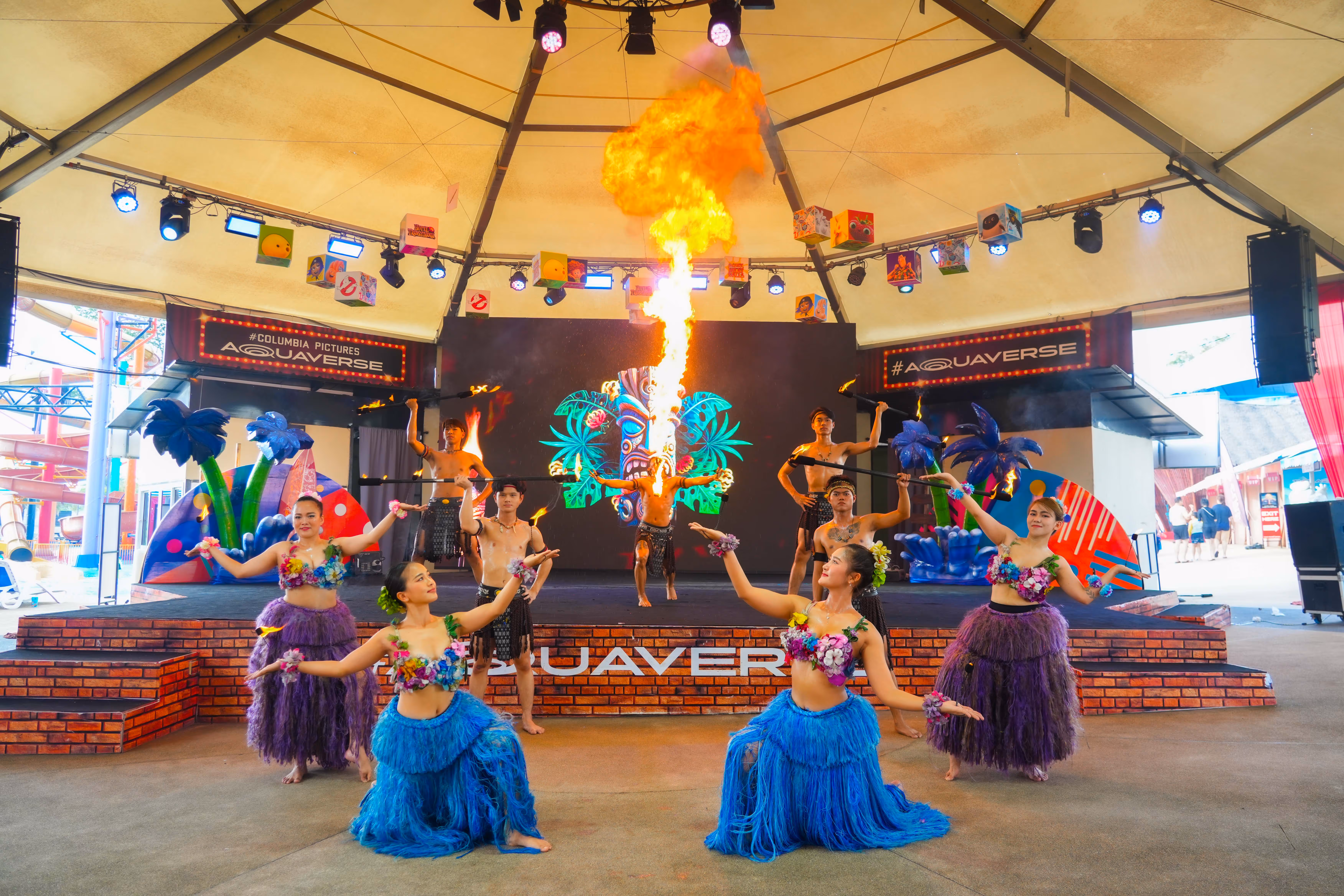 Live performance at Columbia Pictures Aquaverse featuring dancers in colorful island costumes and a fire breather on stage under a tented canopy, with tropical decorations and vibrant lighting.

