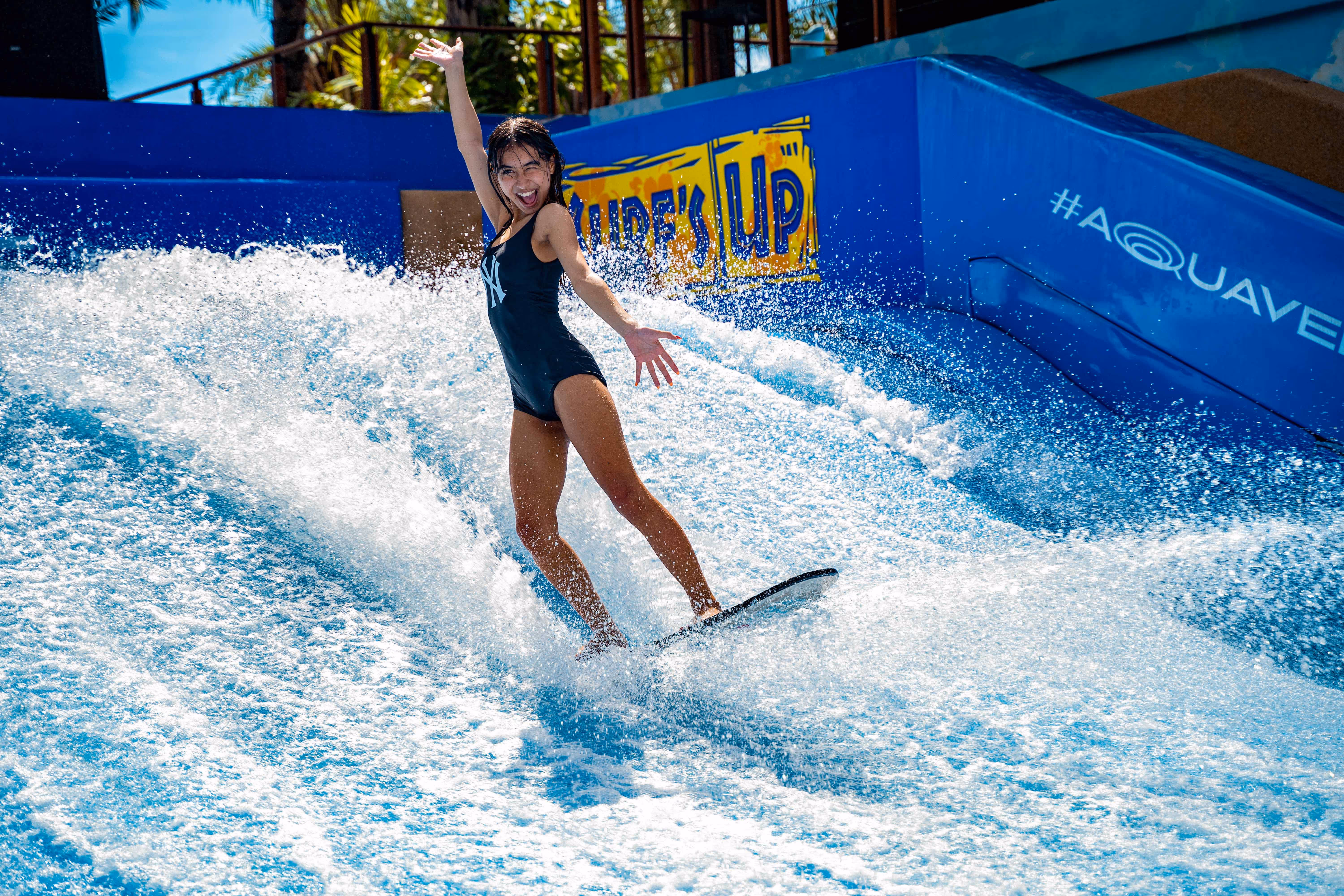 Woman surfing on a flow rider at Columbia Pictures Aquaverse’s Surf’s Up attraction, striking a fun pose with water splashing around and branded signage in the background.

