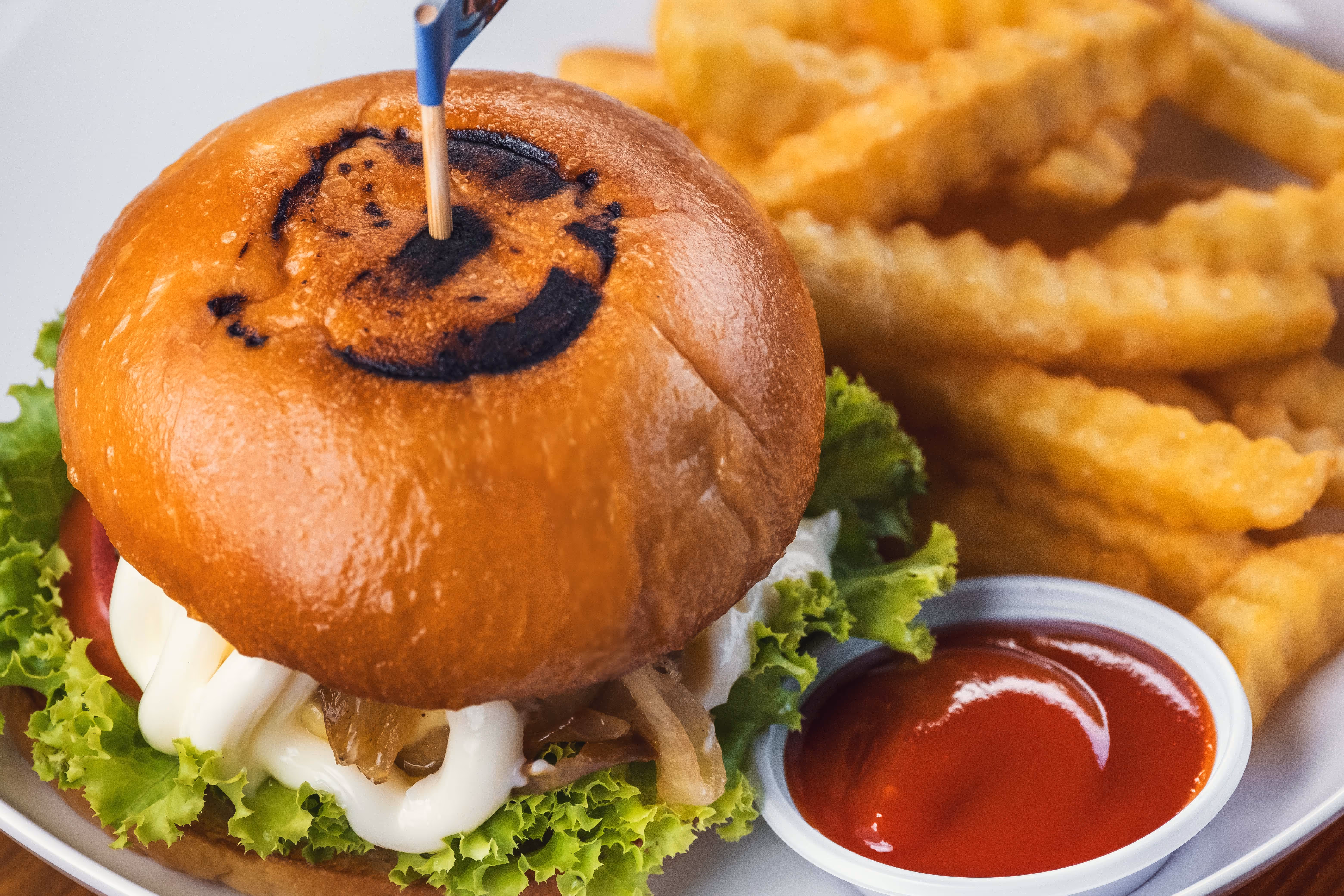 Close-up of a crispy chicken sandwich with lettuce, mayonnaise, and a branded toasted bun featuring the Jumanji logo, served with crinkle-cut fries and a side of ketchup.

