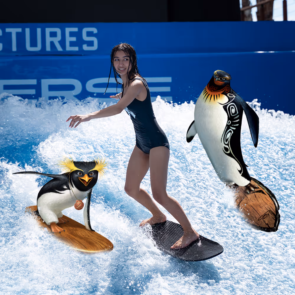 Young woman surfing on an artificial wave alongside animated penguin characters from Surf’s Up, with a vibrant Aquaverse backdrop.

