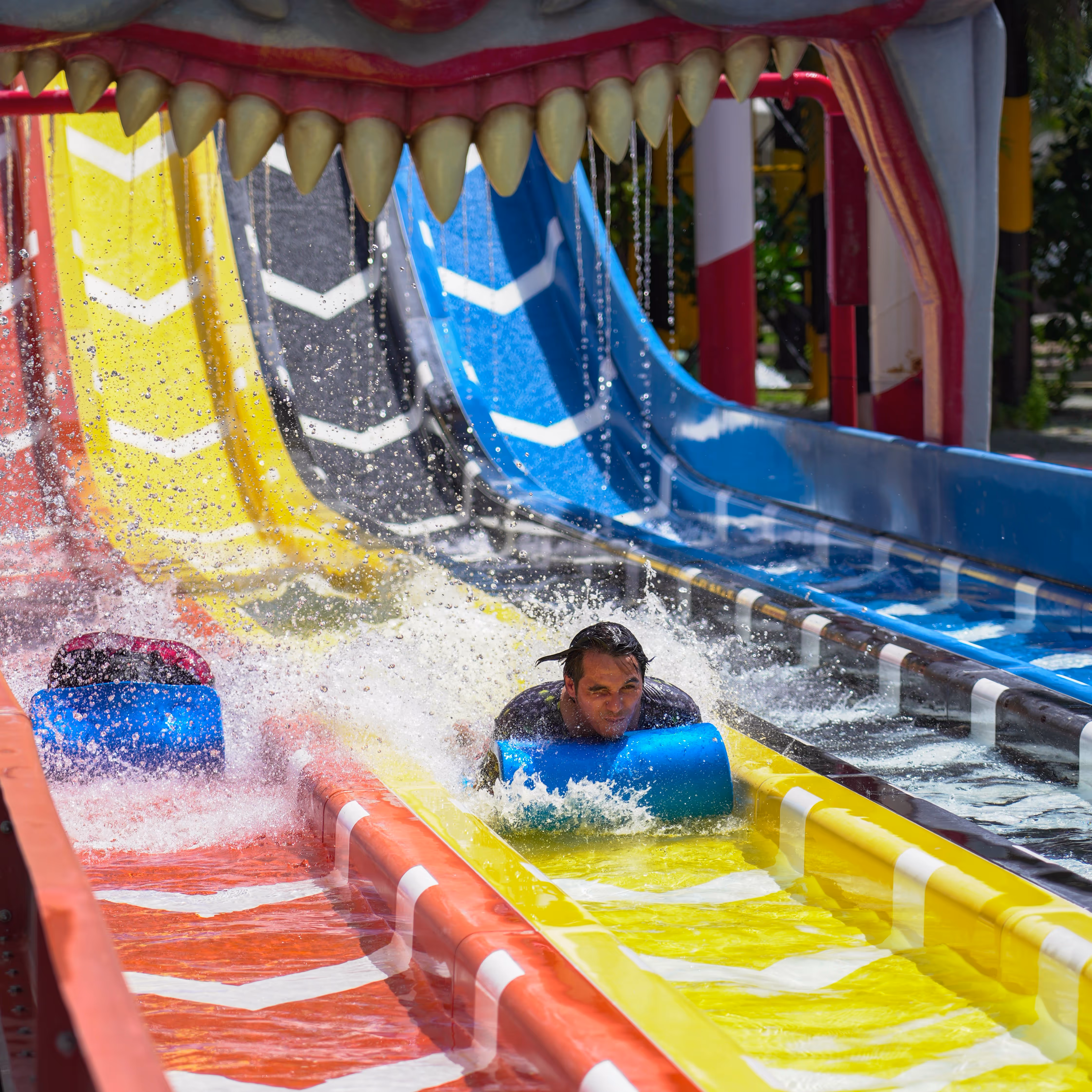 Guest racing down a colorful multi-lane water slide at Columbia Pictures Aquaverse, splashing into the pool beneath a giant clown mouth structure as part of the Zombieland-themed attraction.

