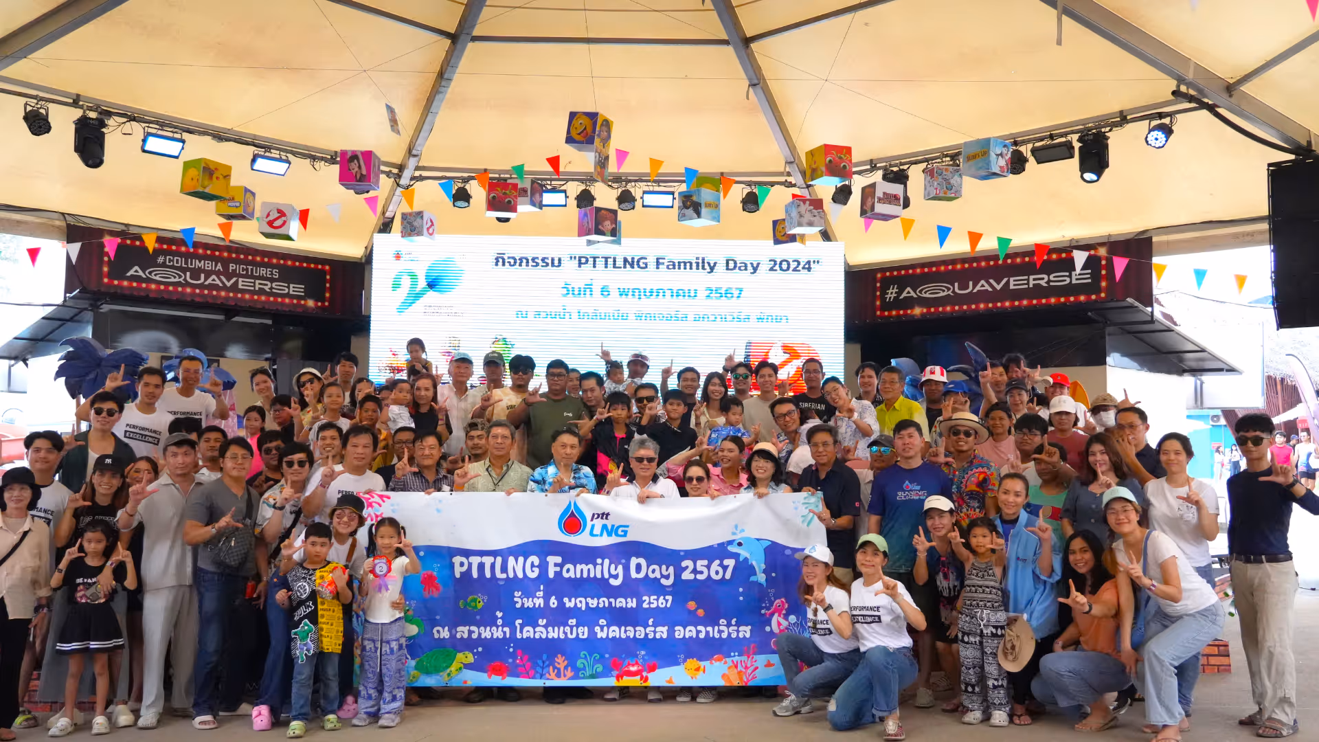 A large group of families and individuals pose together on an indoor stage beneath a tented roof for a commemorative photo during “PTTLNG Family Day 2024.” A banner held at the front reads the event title, date, and venue, with colorful illustrations of water park elements. The background features a large screen with welcoming text and decorations like mini flags and cartoon images hanging above. The crowd is cheerful, many smiling and making peace signs, celebrating a fun-filled corporate family event at Columbia Pictures Aquaverse.