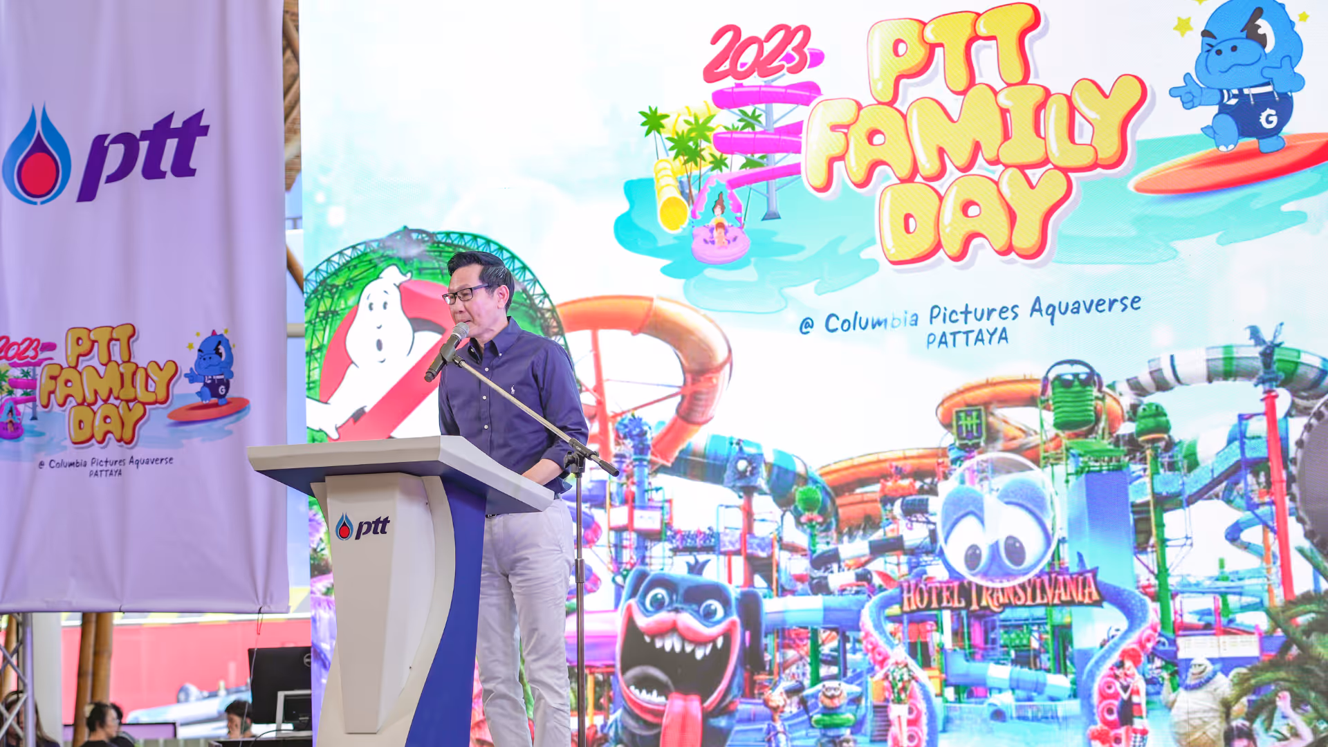 A man in formal attire stands at a podium delivering a speech during the 2023 PTT Family Day event at Columbia Pictures Aquaverse. Behind him, a large, colorful screen displays the event’s branding, including vibrant water slides and cartoon characters from the Hotel Transylvania attraction. Large PTT banners flank the stage, emphasizing the corporate sponsorship. The setting is bright and festive, capturing the excitement of the event’s opening ceremony or keynote moment.