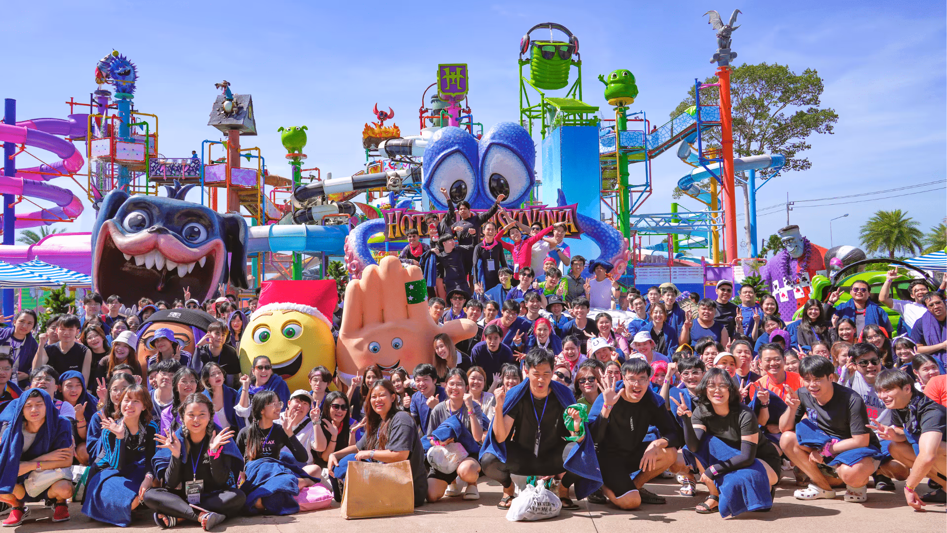 A large, joyful group of students poses in front of the vibrant Hotel Transylvania themed water park zone at Columbia Pictures Aquaverse. Surrounded by giant, colorful cartoon character statues, water slides, and playful structures, the group fills the foreground—many smiling, waving, and flashing peace signs. Mascots and oversized props, including a giant hand and monster faces, enhance the fun and festive atmosphere under a bright blue sky. The image captures the energy and excitement of a school field trip or youth group outing.