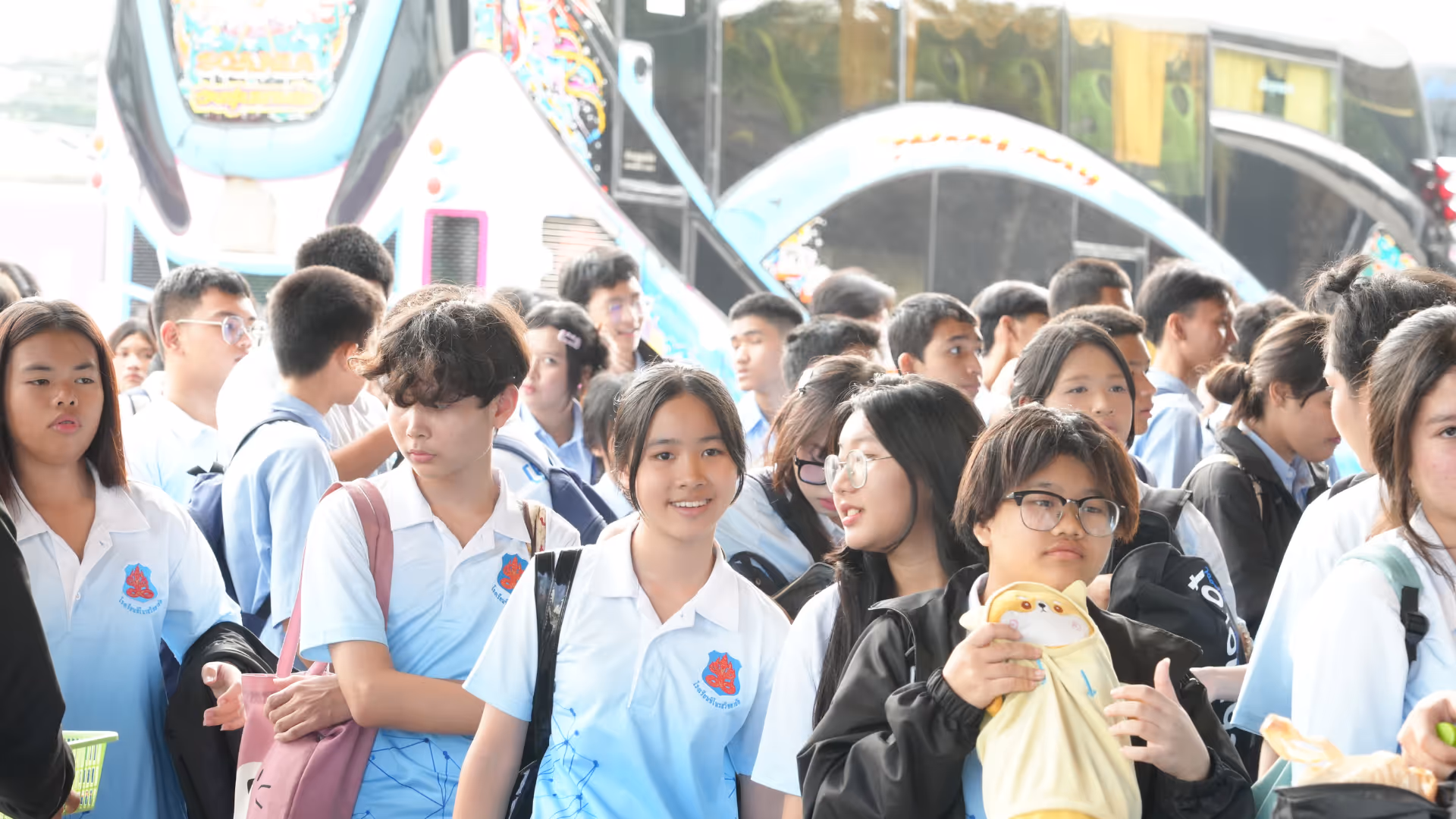 A large group of students, mostly wearing light blue and white school uniforms with a red emblem, gathers in a lively area of what appears to be a theme park. Some students carry snacks or drinks, while others chat and smile. In the background, colorful attractions and signage suggest a fun and energetic environment, possibly during a school field trip or educational outing. The overall mood is bright, social, and filled with youthful excitement.