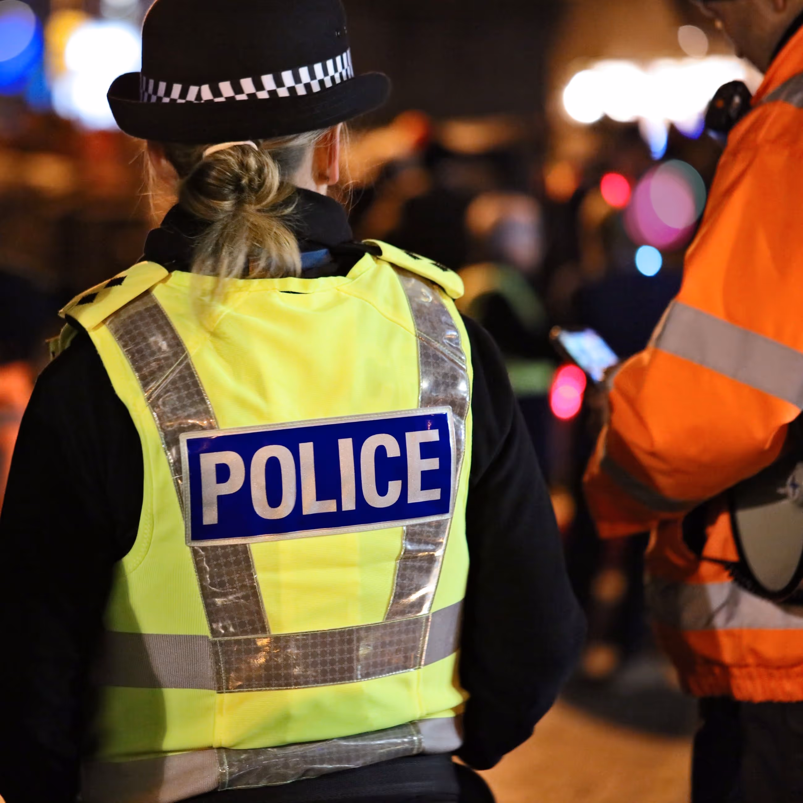 A policewoman seen from the back, wearing a yellow 'POLICE' vest and a black hat.