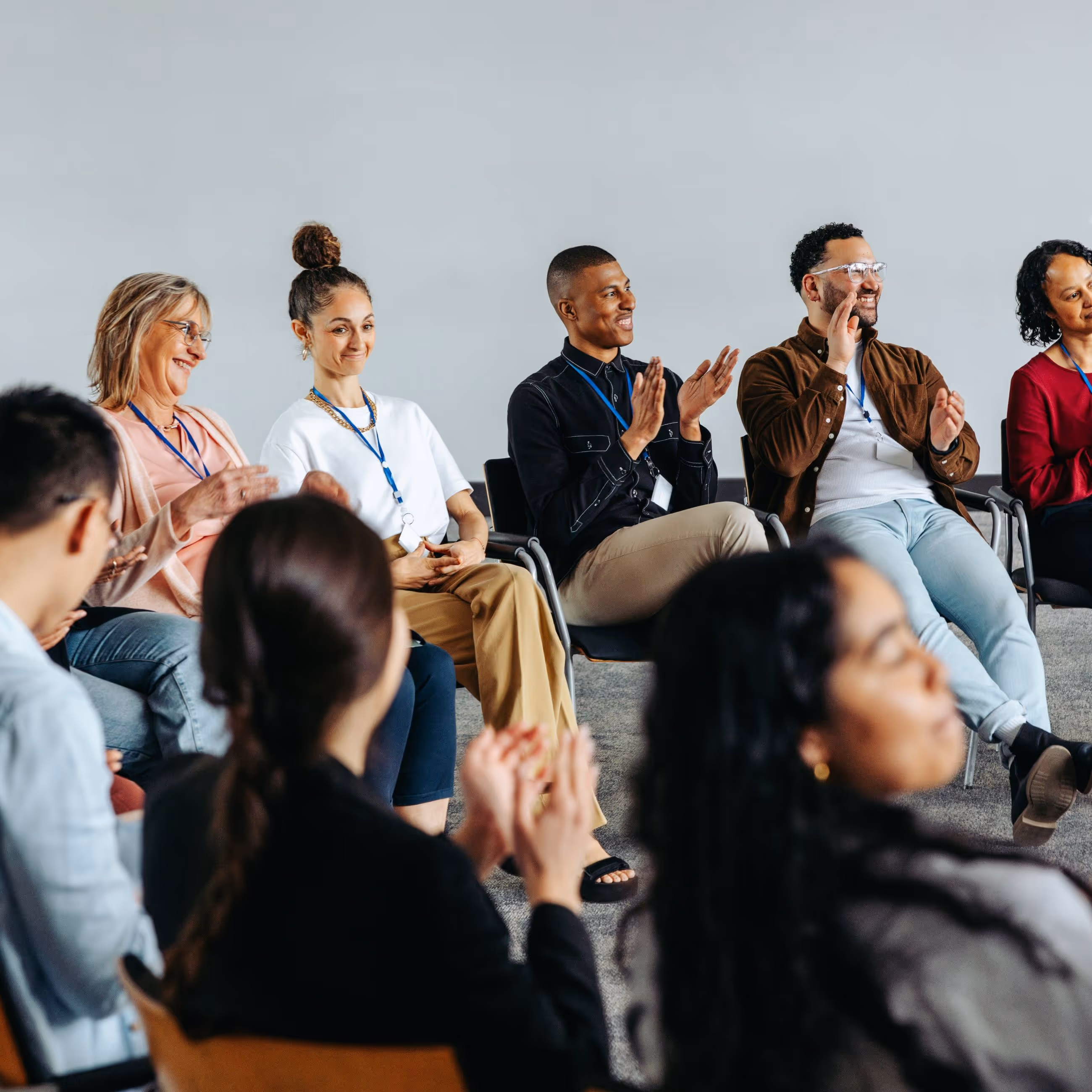 A diverse group of people sit in a semicircle of chairs, listening to one of the men in the circle speaking