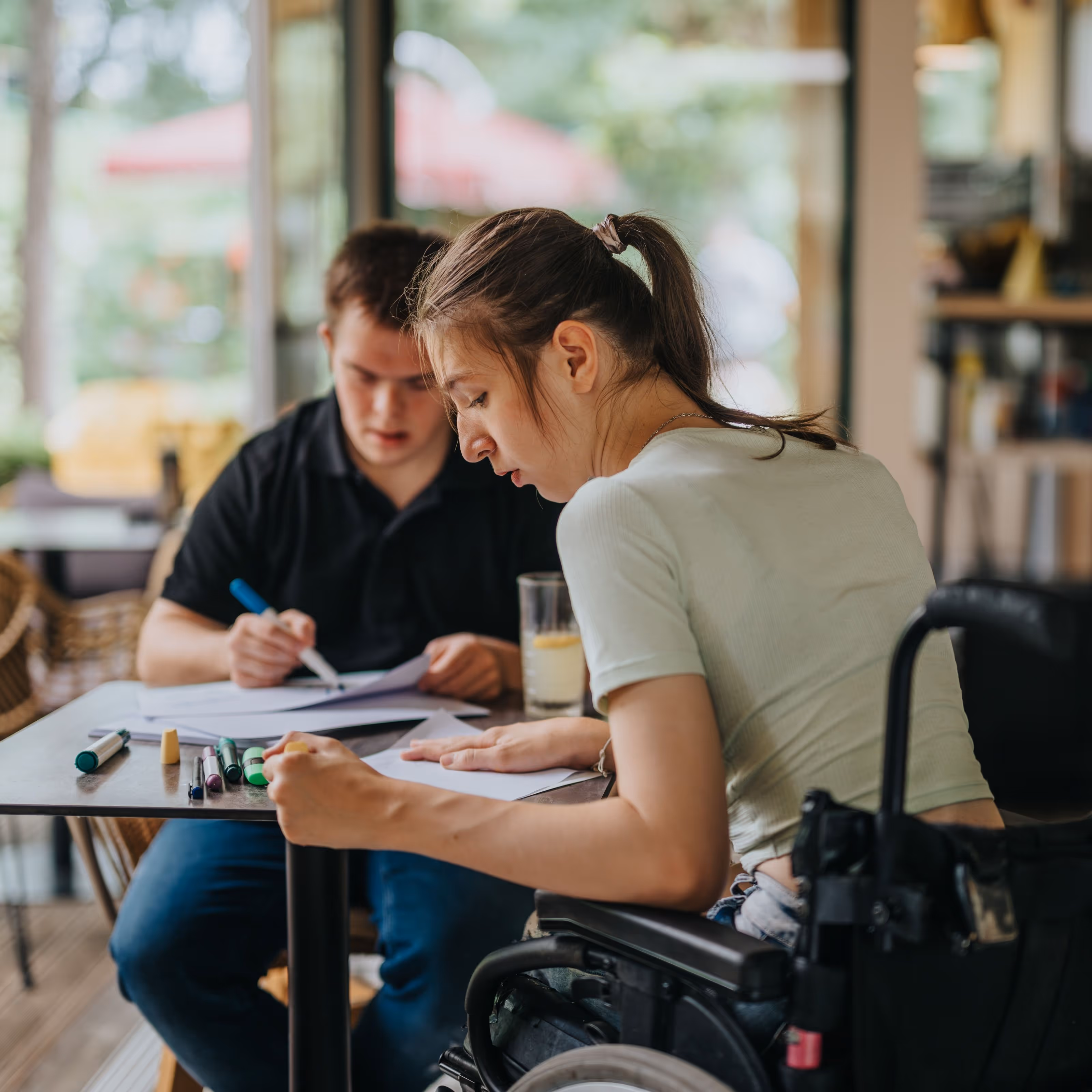 Two young people, one a wheelchair user, sit at a desk studying.