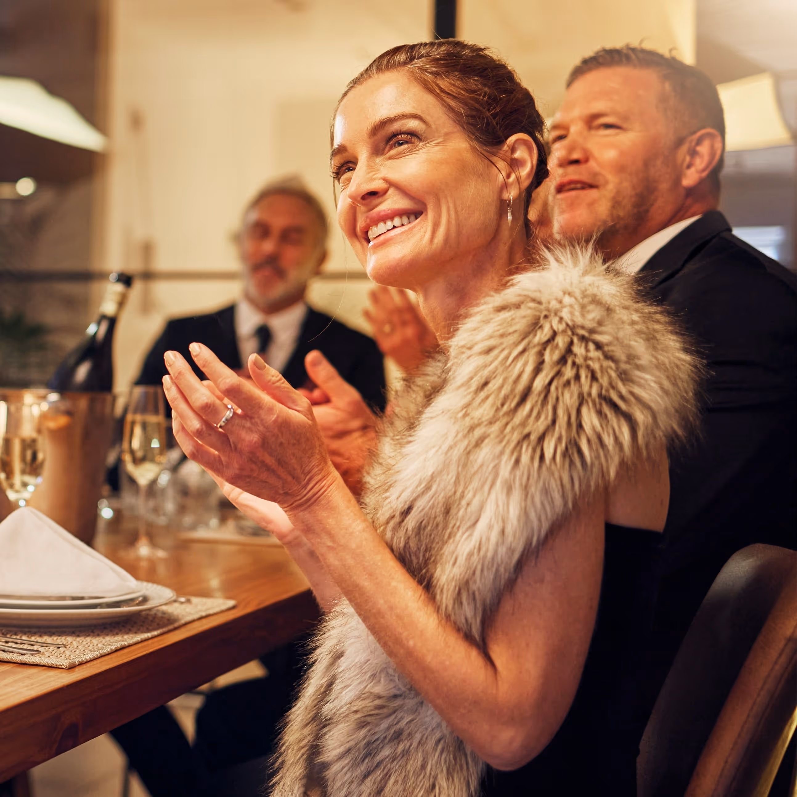 A woman in formal evening dress sits around a long dining table clapping to mark the end of a speech.